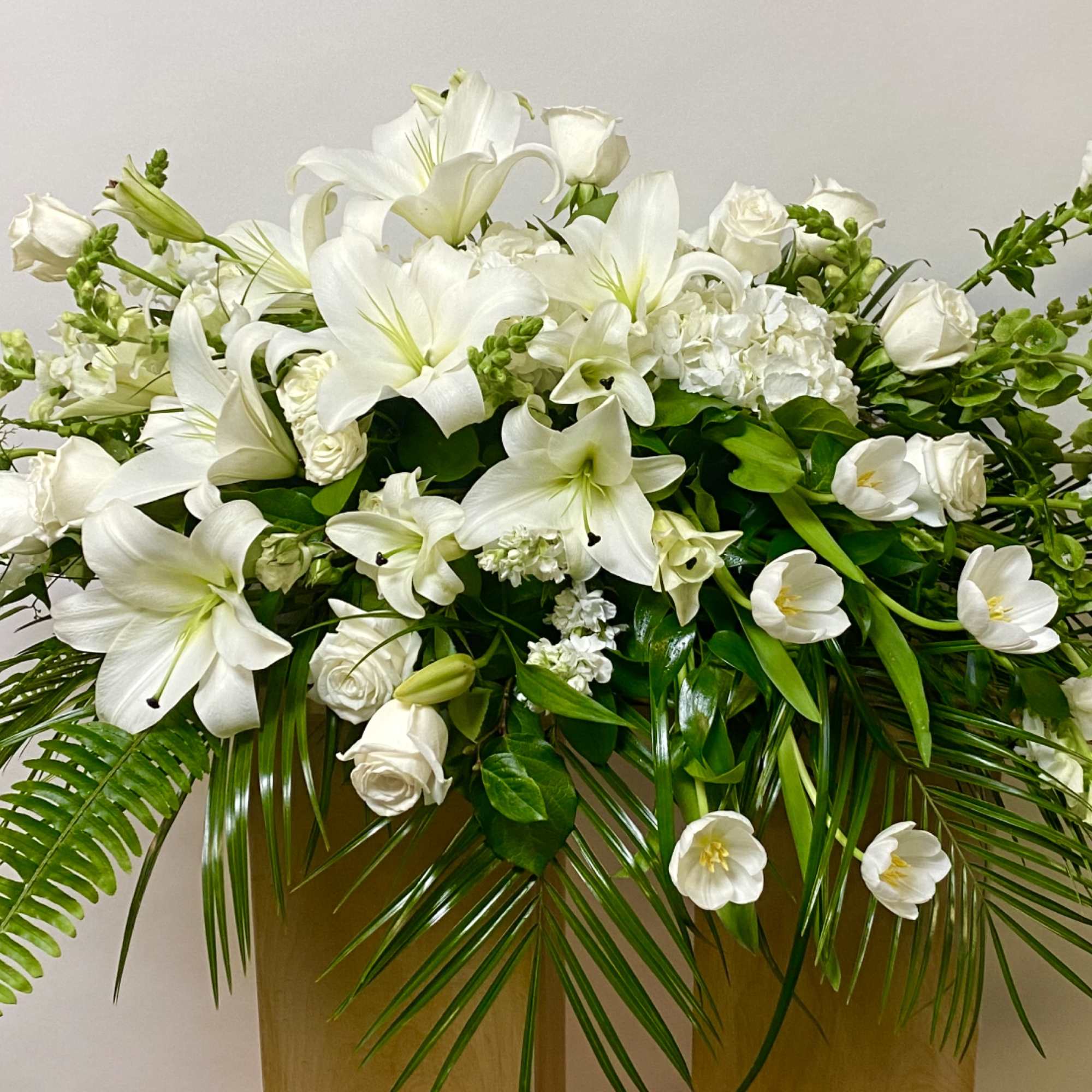 White lilies and roses arranged in a gold container with palm fronds.