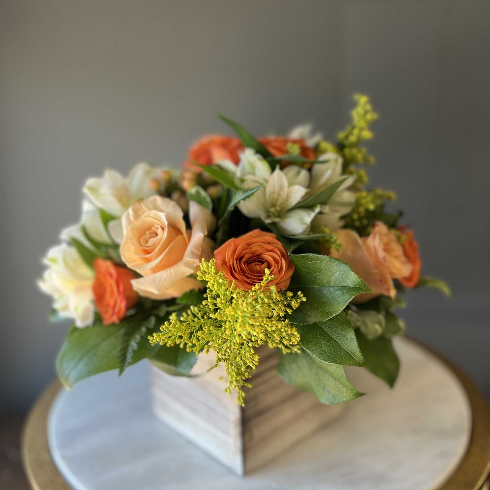 Compact arrangement of orange and peach roses with white blooms in a wooden cube vase.