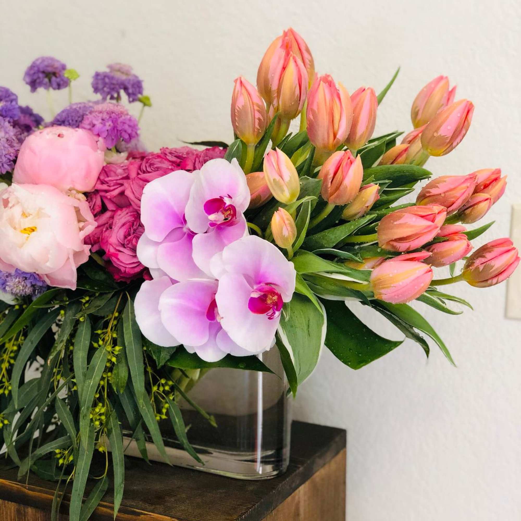 Mixed bouquet with pink orchids, tulips, roses, and purple blooms in a glass vase