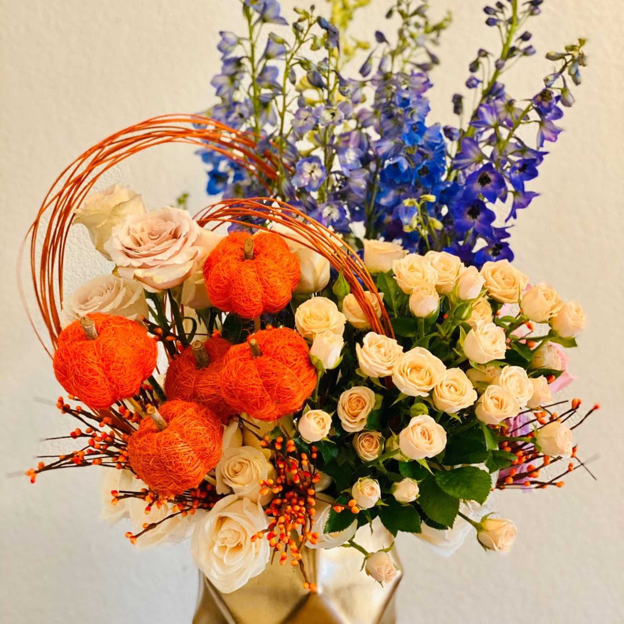 Bouquet of cream roses and blue delphinium with orange pumpkin accents in a gold vase
