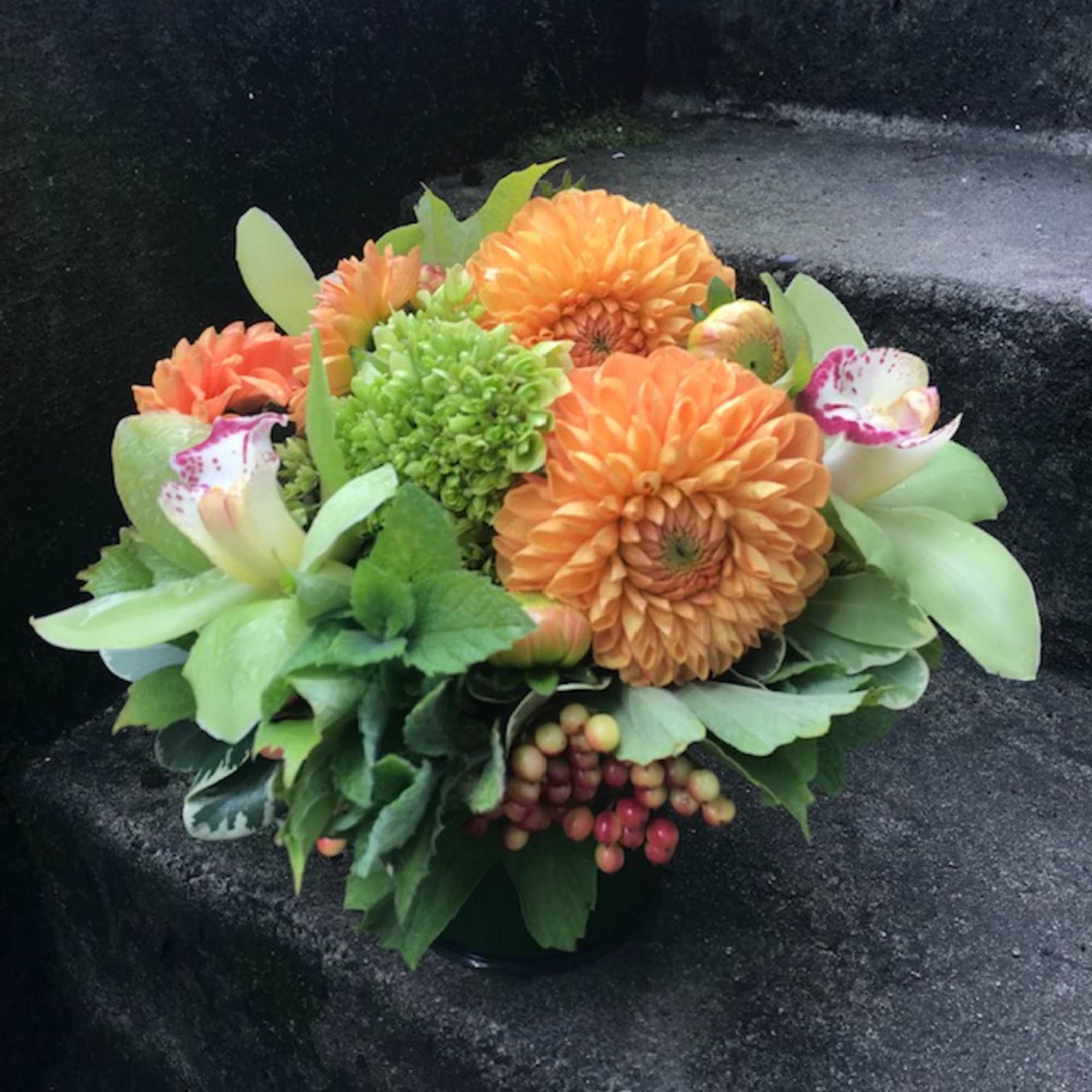 Orange and green floral arrangement with spotted white orchids in a low container