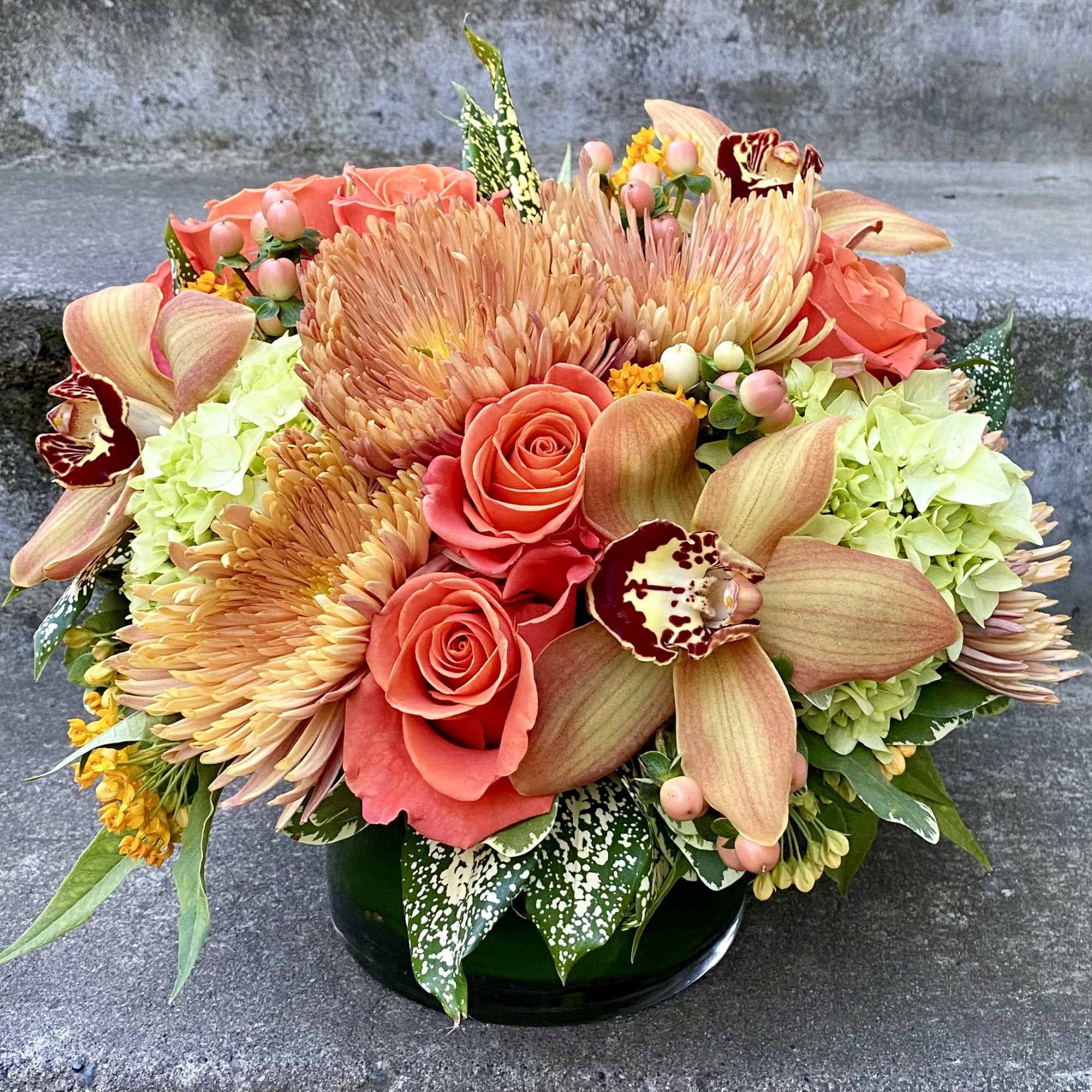 Coral roses and peach orchids arranged with pale hydrangeas in a black vase.