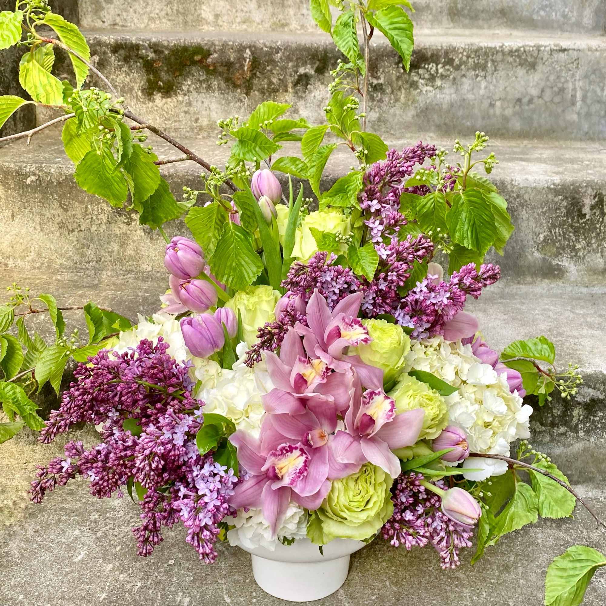 Pink orchids and lilacs arranged with white blooms in a white vase.