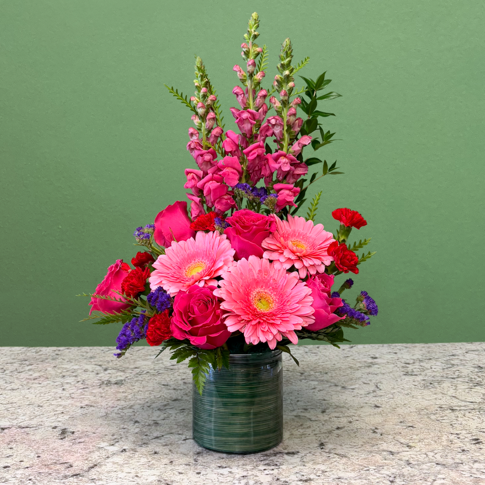 Tall pink floral arrangement with gerbera daisies, roses, and snapdragons in a green glass vase
