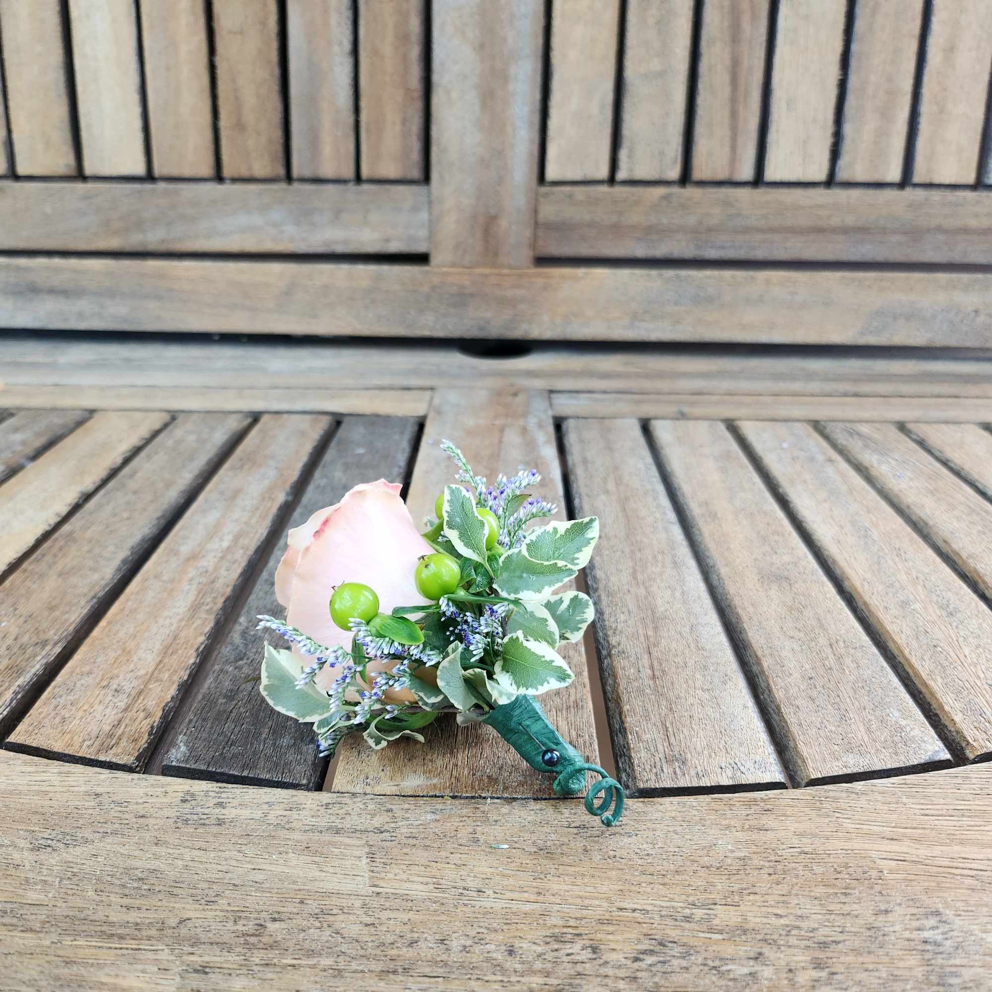 Small boutonniere with a pale pink rose and greenery on a wooden bench
