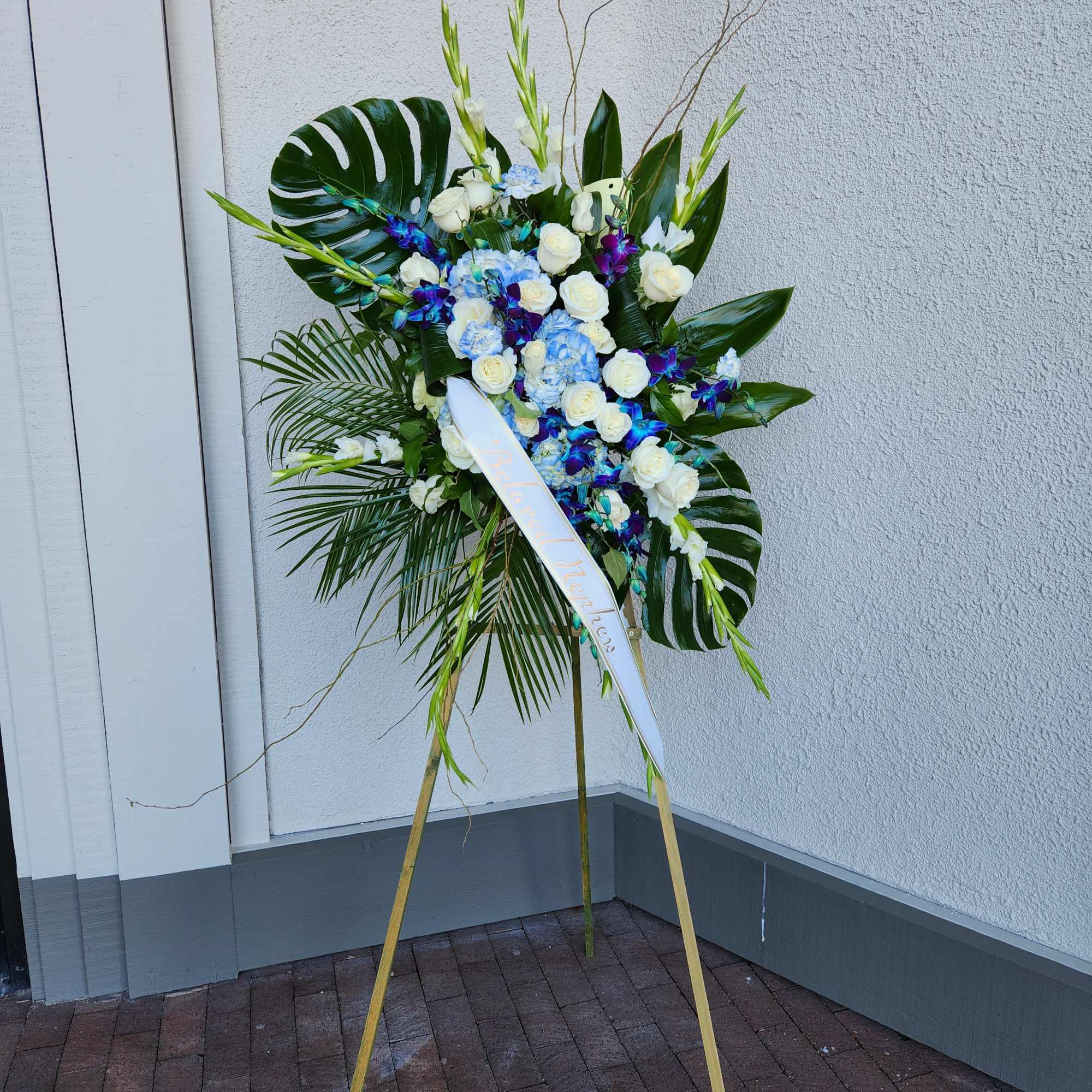 Tall standing floral spray with white roses, blue hydrangeas, and tropical leaves on an easel