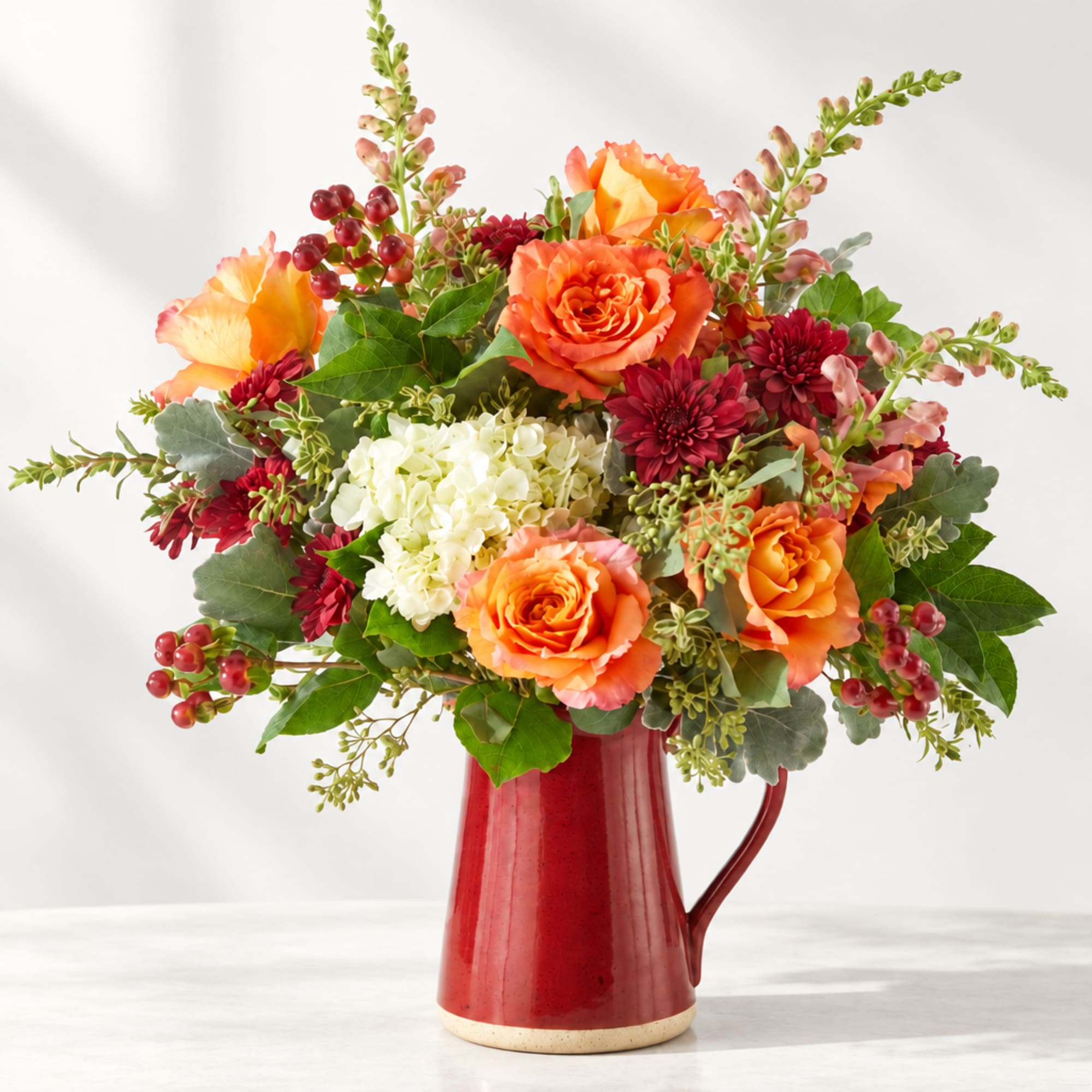 Orange and burgundy flower arrangement in a red pitcher vase