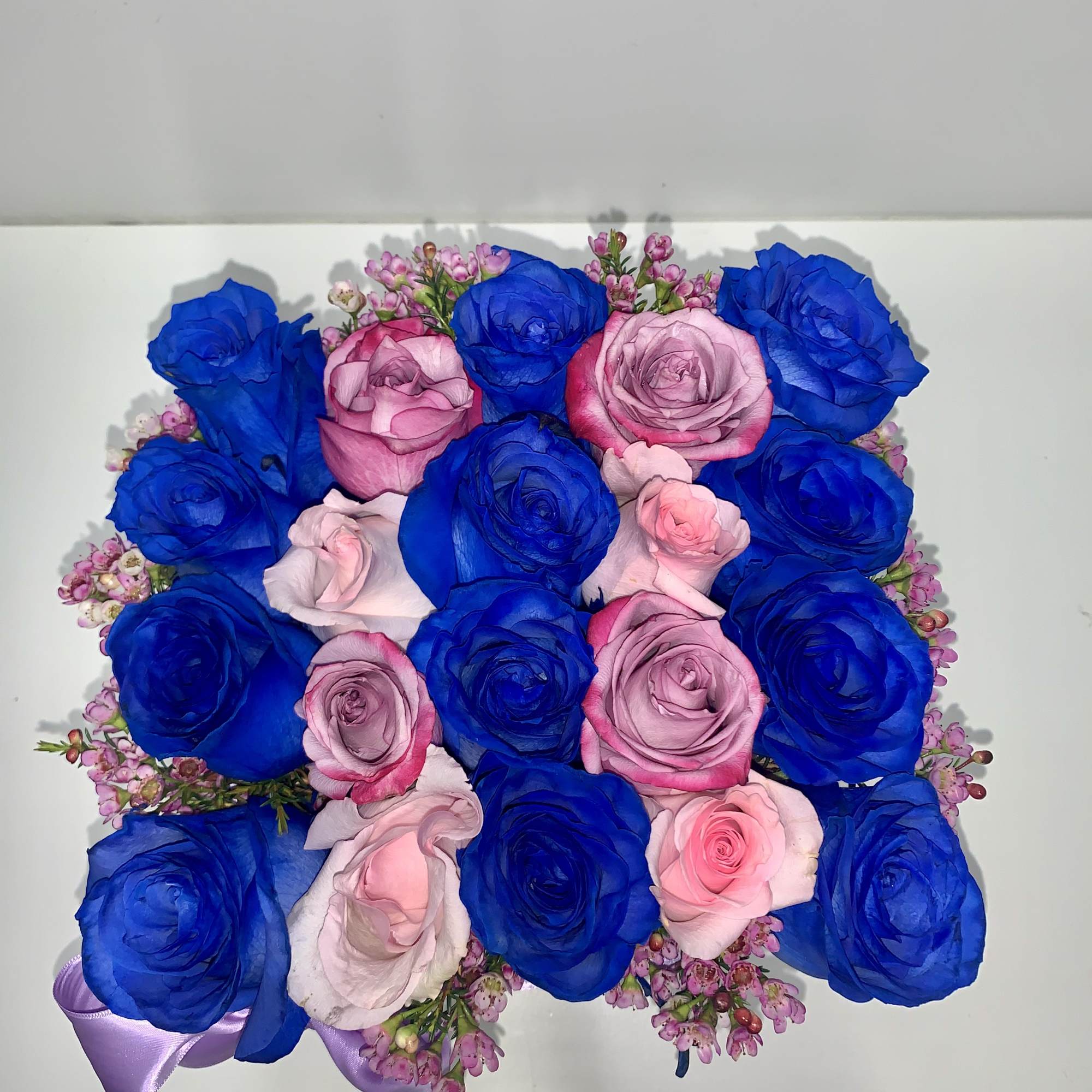 Square arrangement of vivid blue and soft pink roses topped with tiny pink blooms and a lavender ribbon accent.