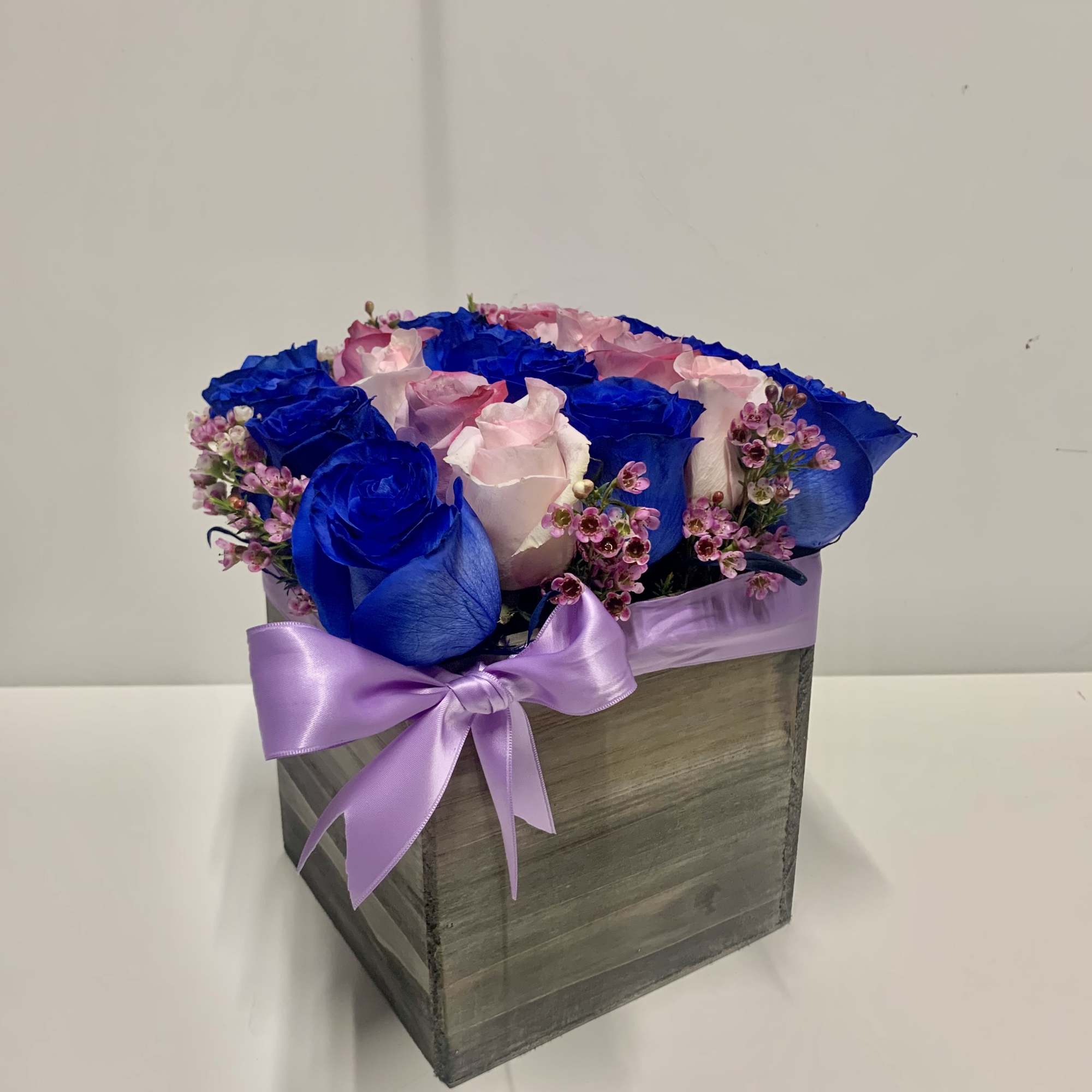 Square wooden box of blue and light pink roses with tiny pink blooms and a lavender ribbon bow