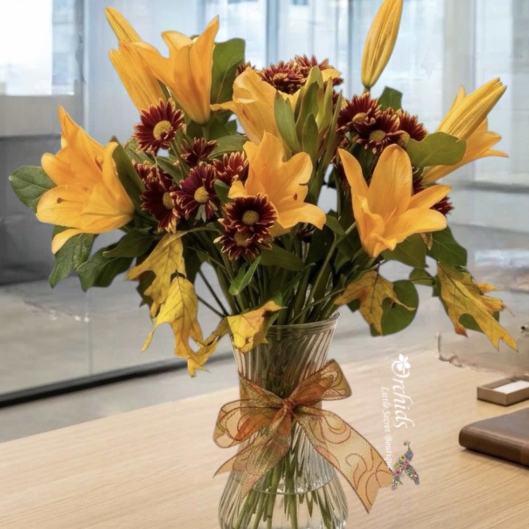 Orange lilies and burgundy daisies in a clear glass vase with a ribbon