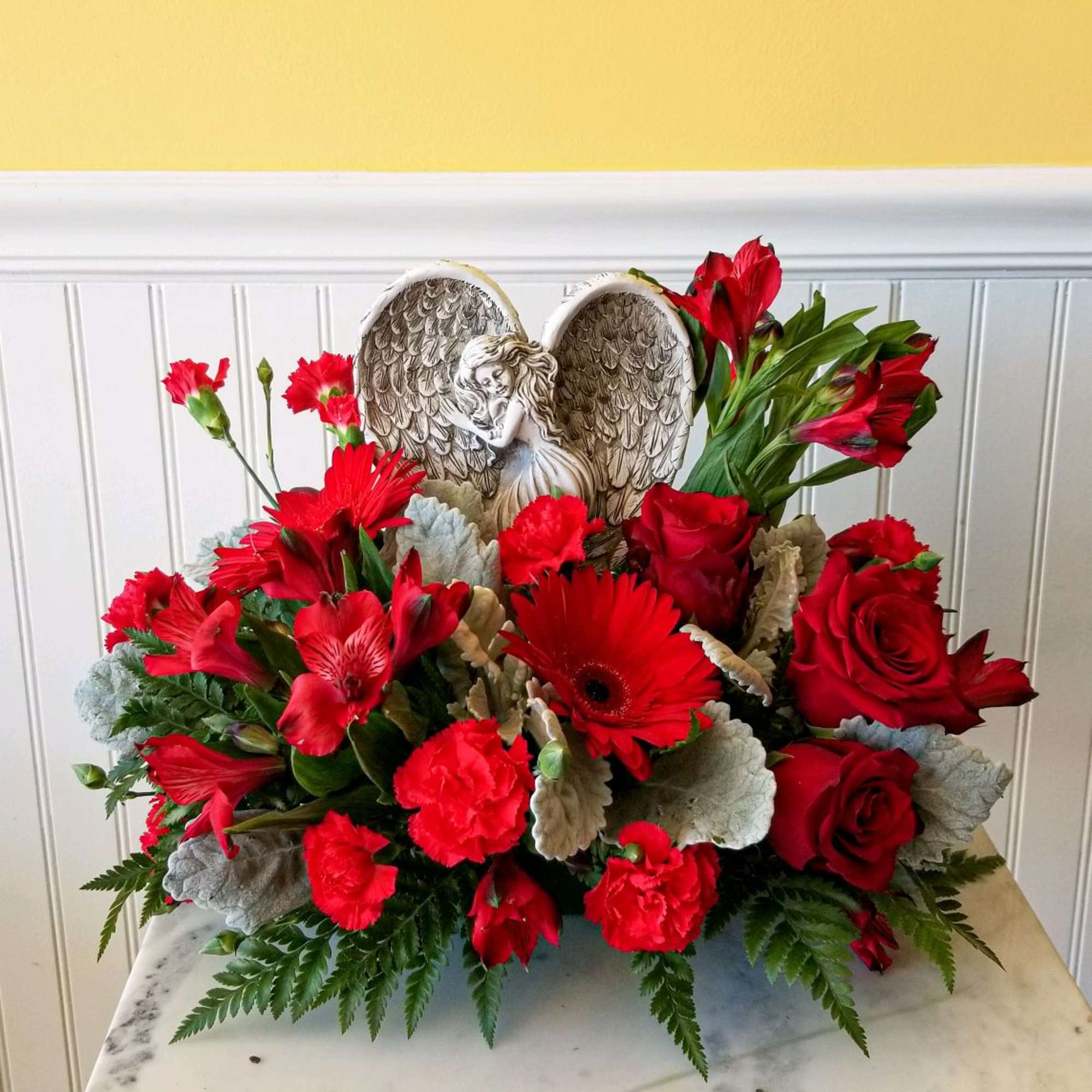 Red floral arrangement with roses, gerbera daisies, and carnations around an angel wing ornament