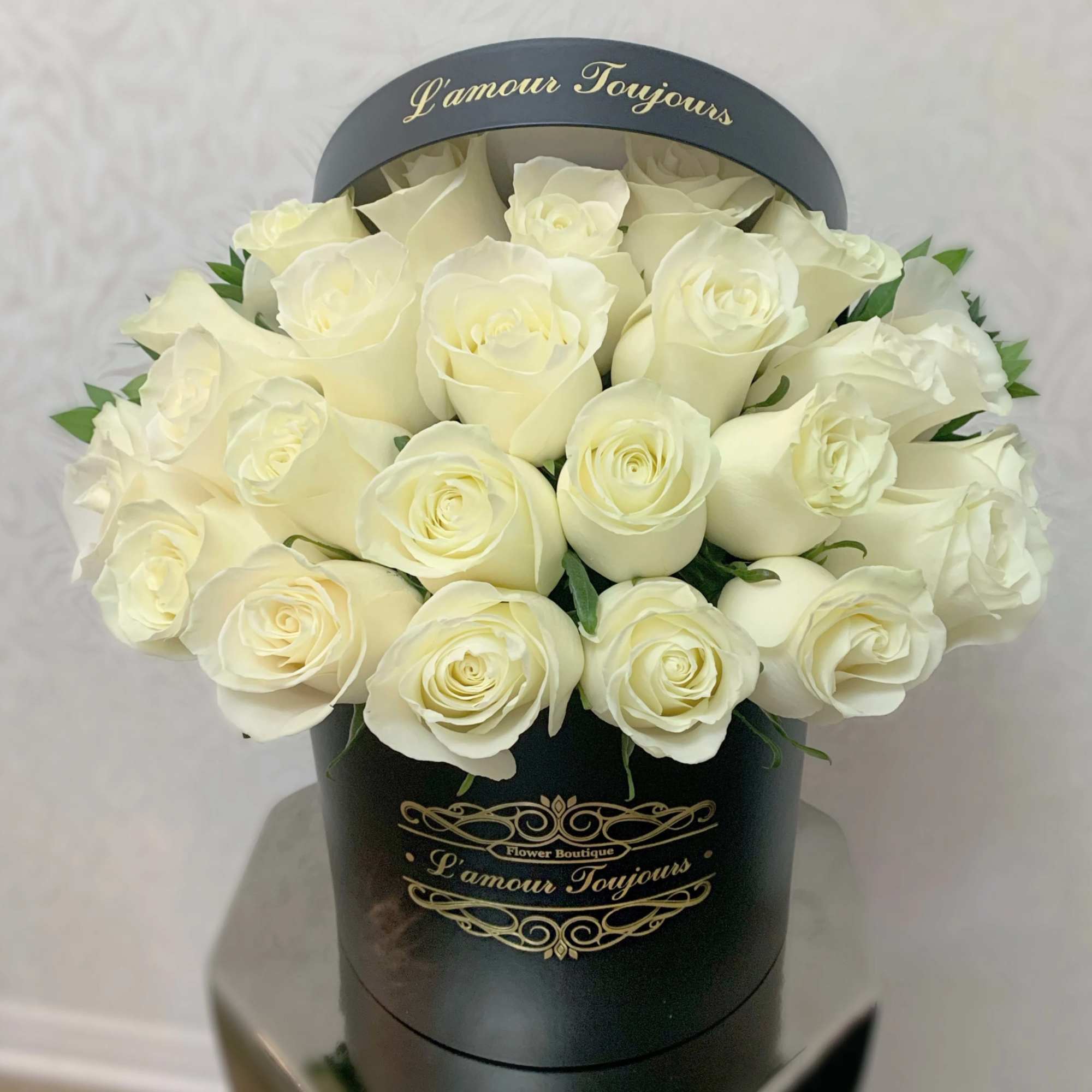Round black hatbox filled with tightly packed ivory roses with gold script text.