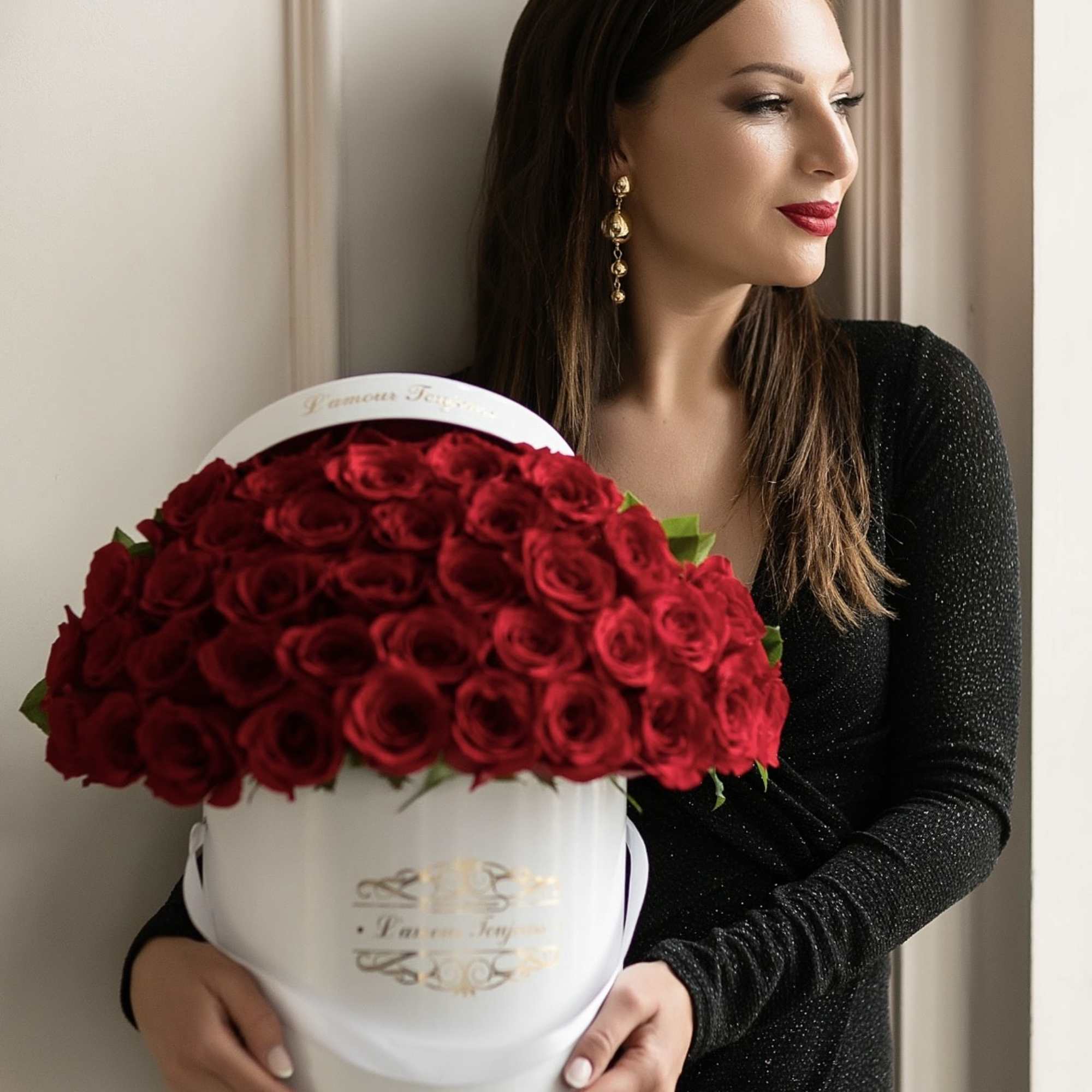 Woman in a black dress holding a round white box filled with dozens of red roses.