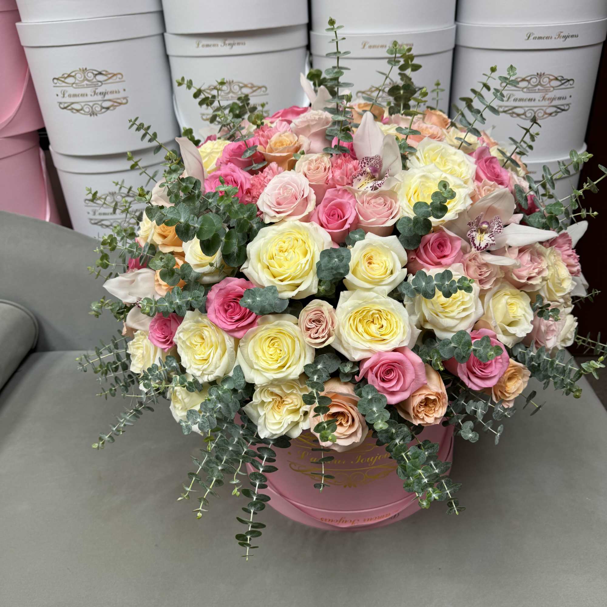 Round hat box arrangement of pink, yellow, and cream roses with orchids and trailing eucalyptus.