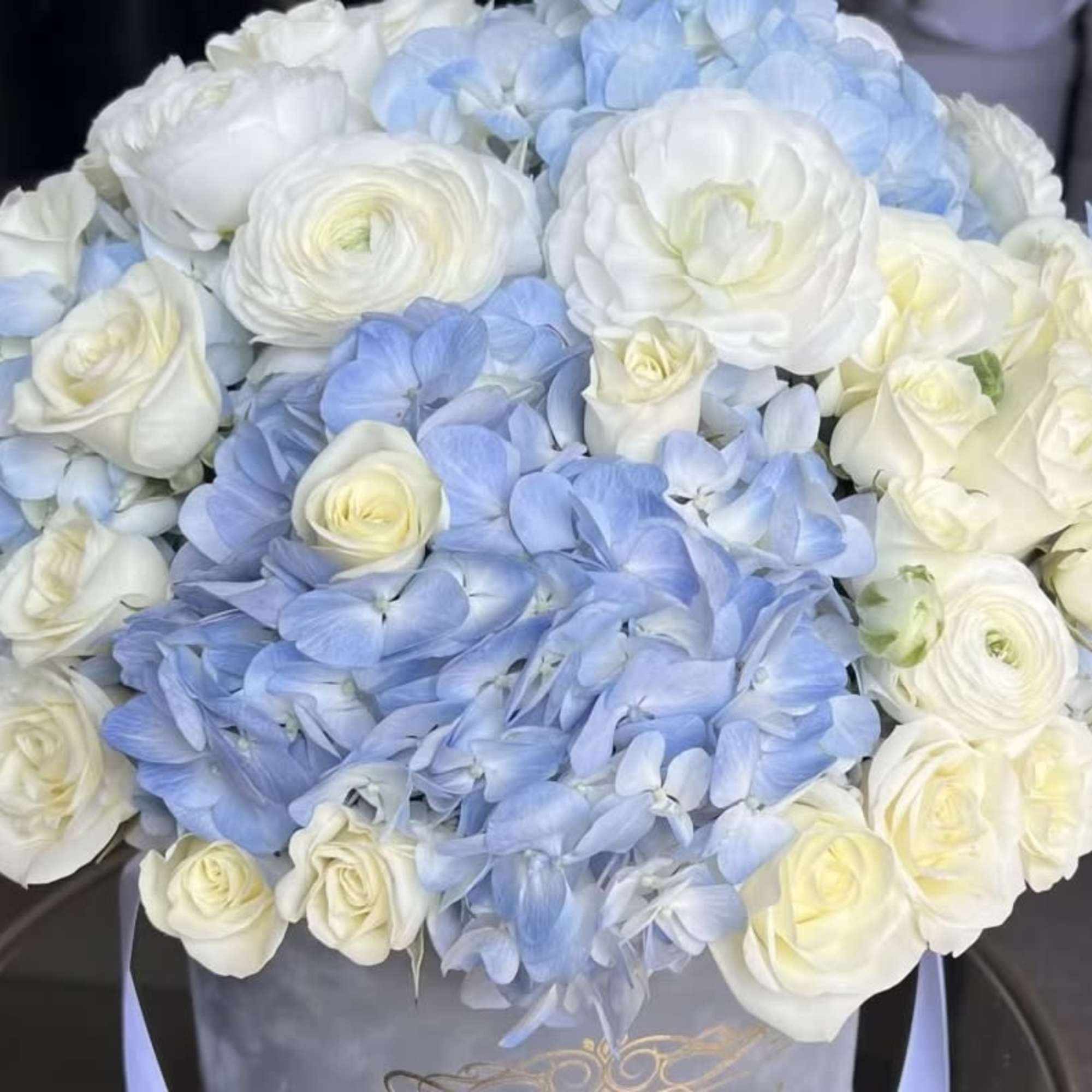 Round arrangement of white roses and ranunculus with light blue hydrangeas in a hatbox-style container