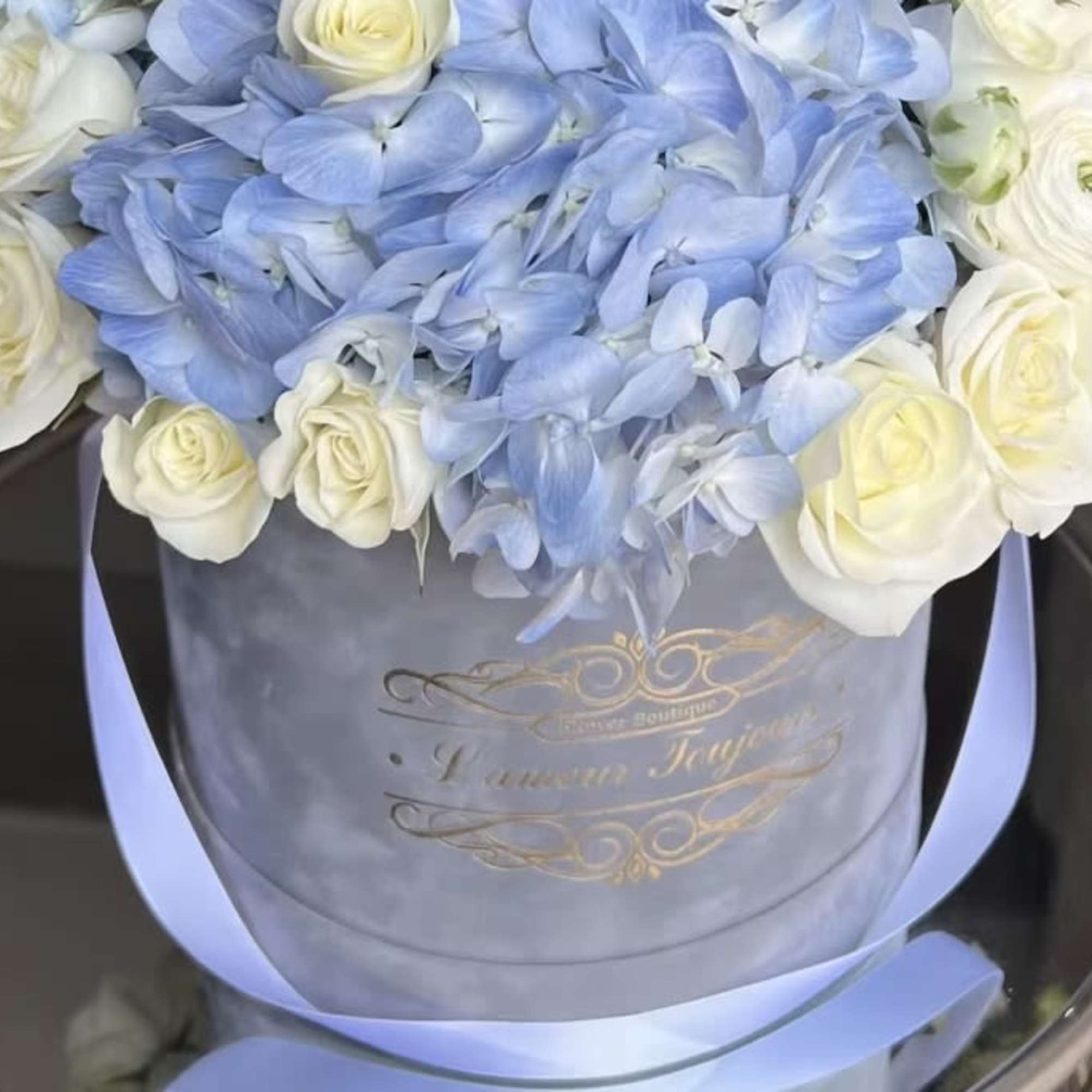 Round hatbox arrangement of pale blue hydrangeas and white roses with a blue ribbon.