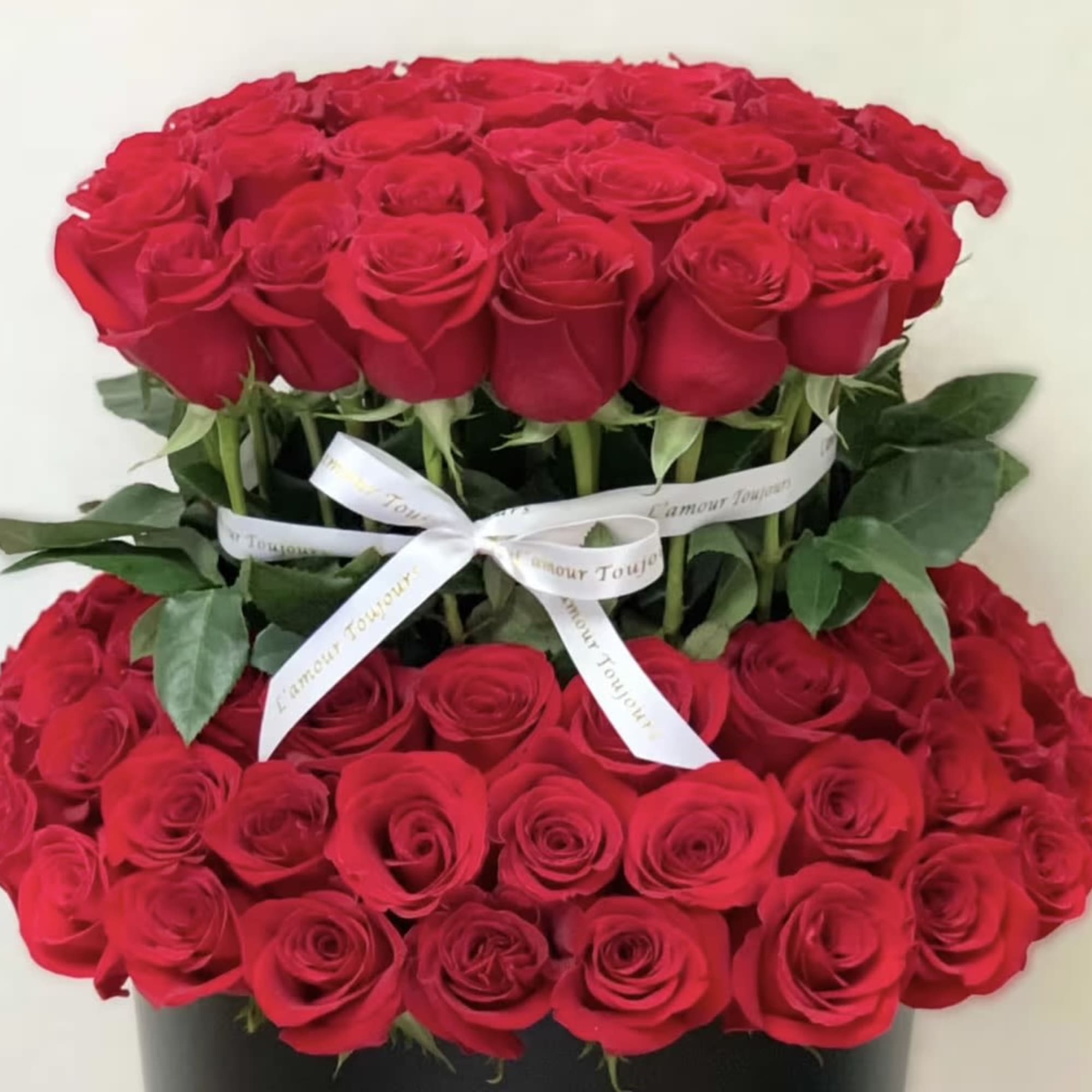 Two-tier arrangement of red roses in a black hatbox with white ribbon bow
