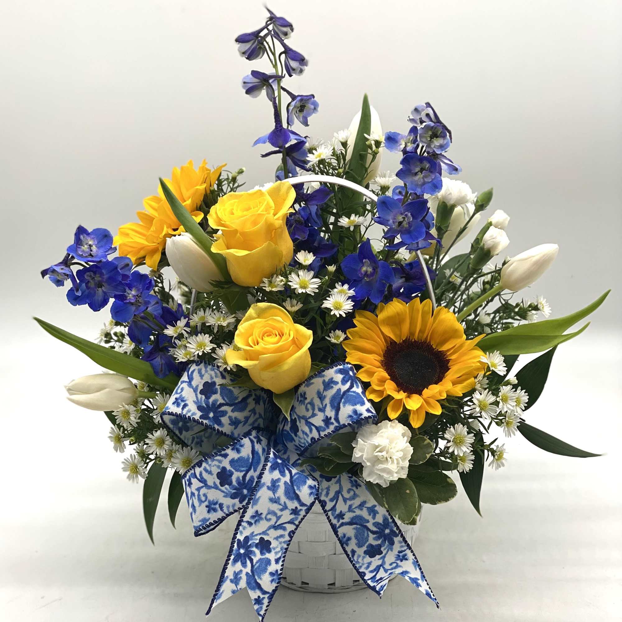Basket arrangement with yellow roses, sunflowers, blue flowers, and white blooms