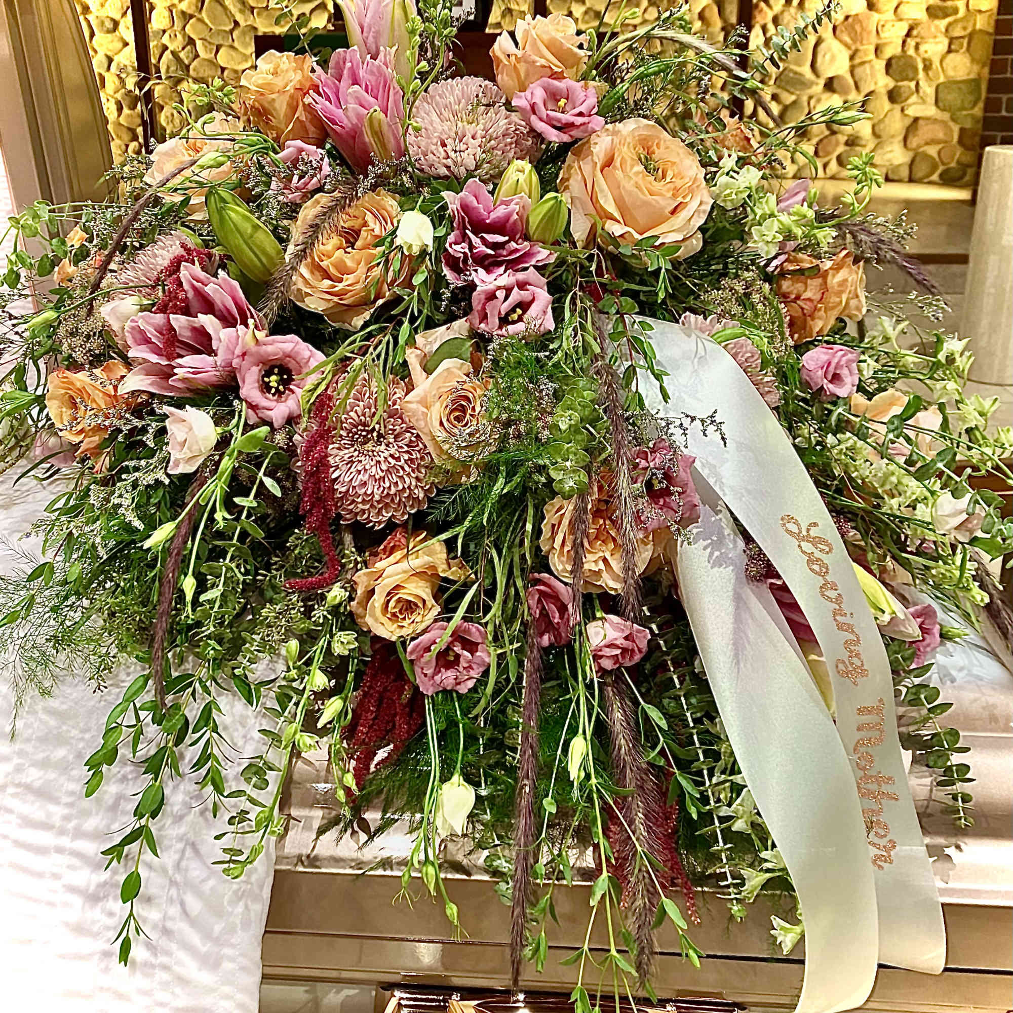 Large peach and pink casket spray with roses, lilies, and mums draping over a bronze casket