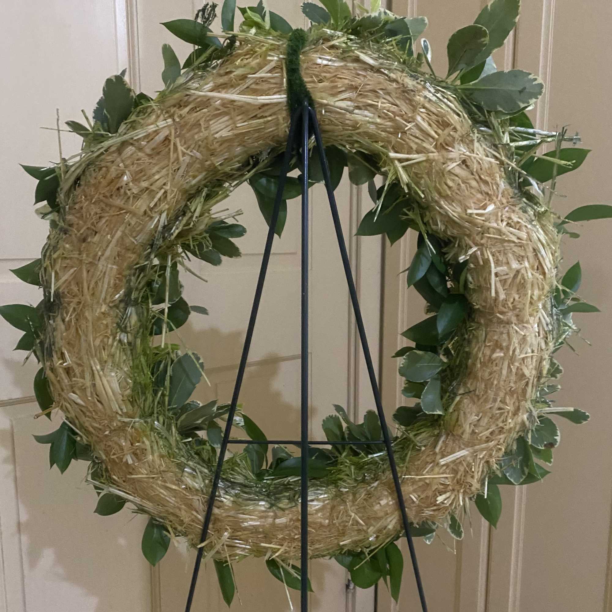 Straw wreath with green foliage on a black metal easel against a neutral wall