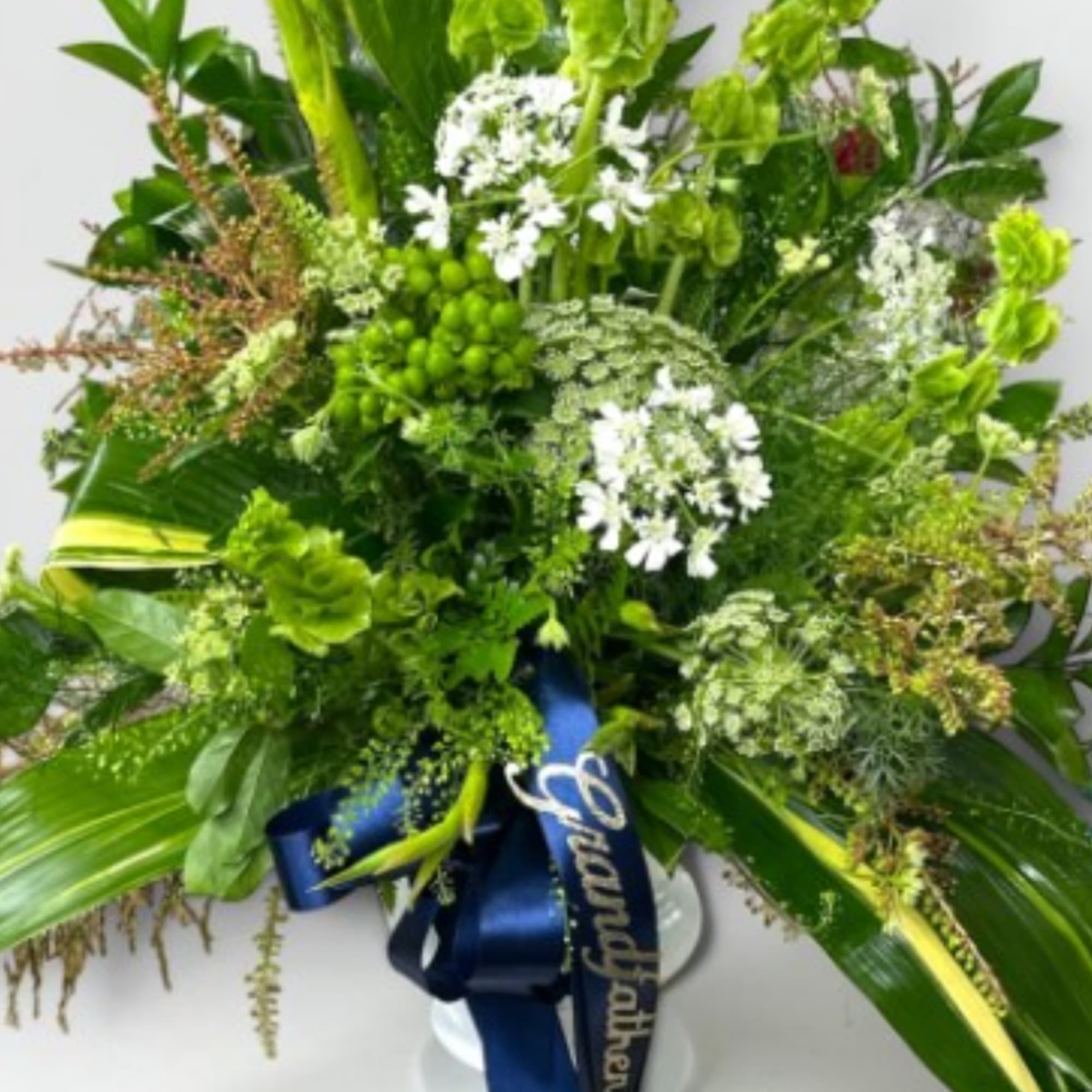 Large green floral arrangement with white blooms and a blue ribbon on a pedestal vase