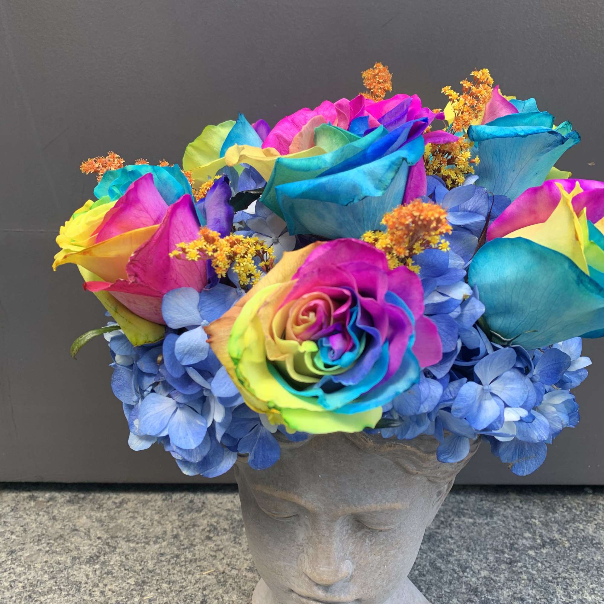 Rainbow roses and blue hydrangeas in a gray bust vase