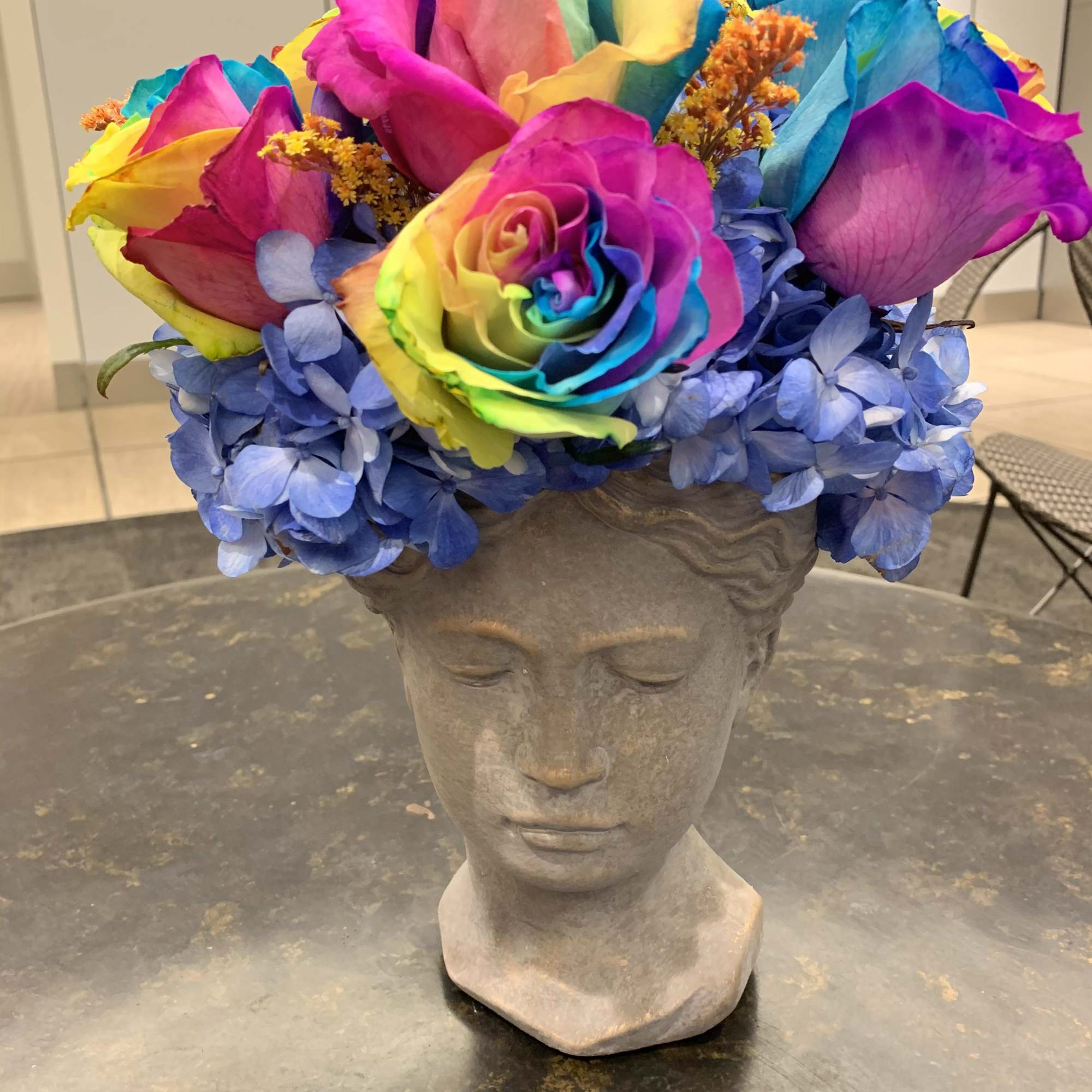 Rainbow rose bouquet in a gray sculpted vase with blue hydrangeas