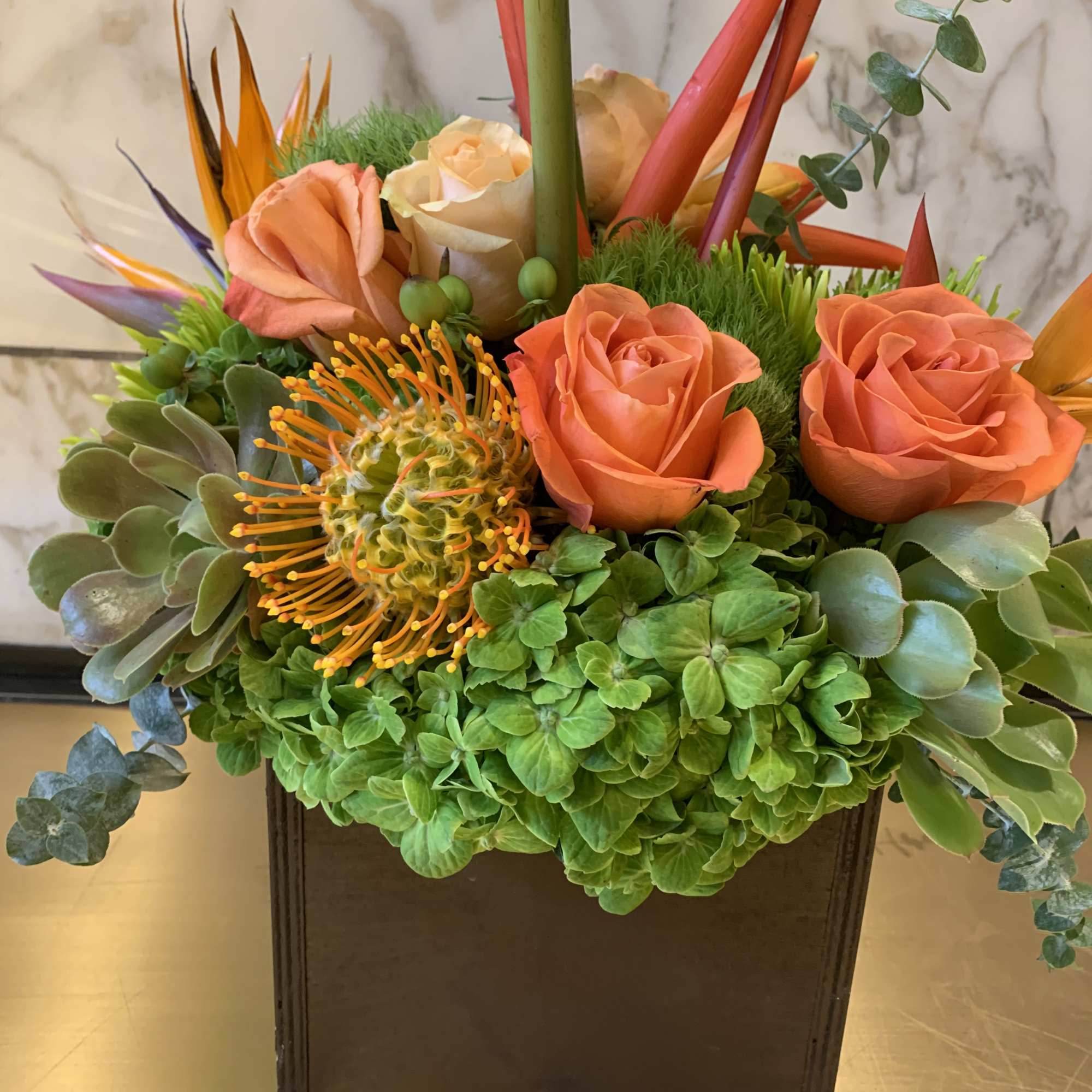 Orange roses in a square black vase with green hydrangea and tropical accents