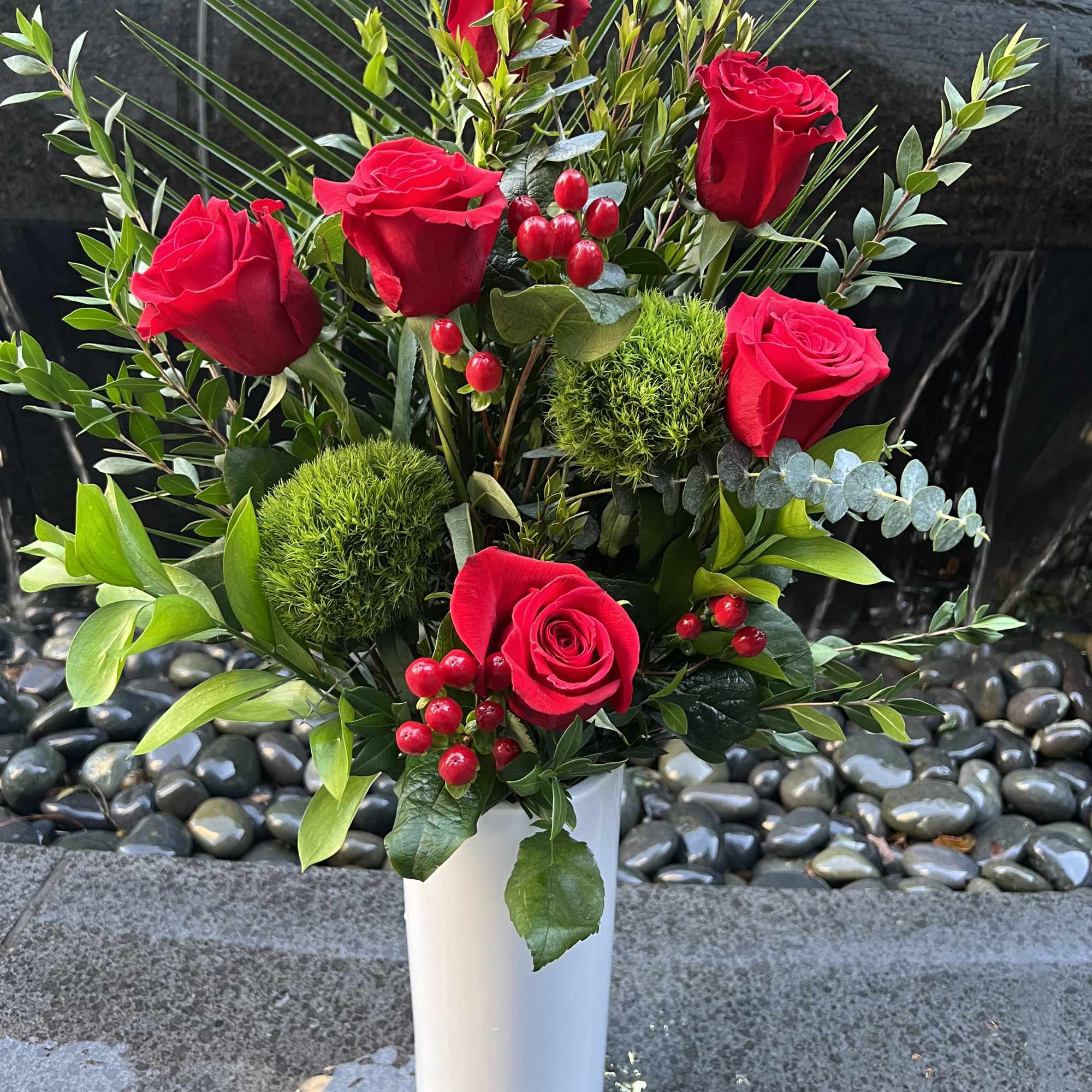 Red roses arranged in a white vase with mixed greenery and berry accents