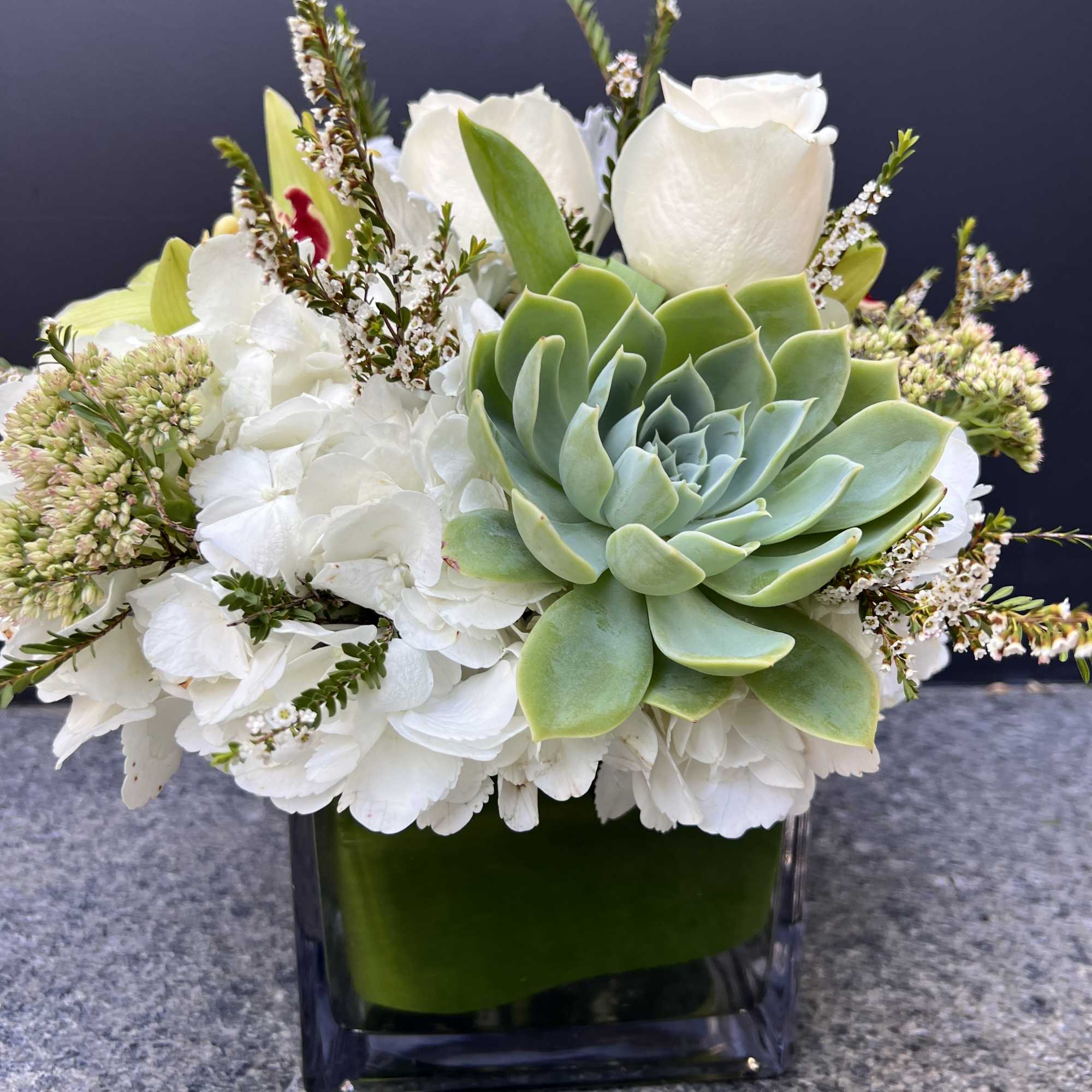 Low white and green arrangement with hydrangeas, roses, a succulent, and small blooms in a glass cube vase