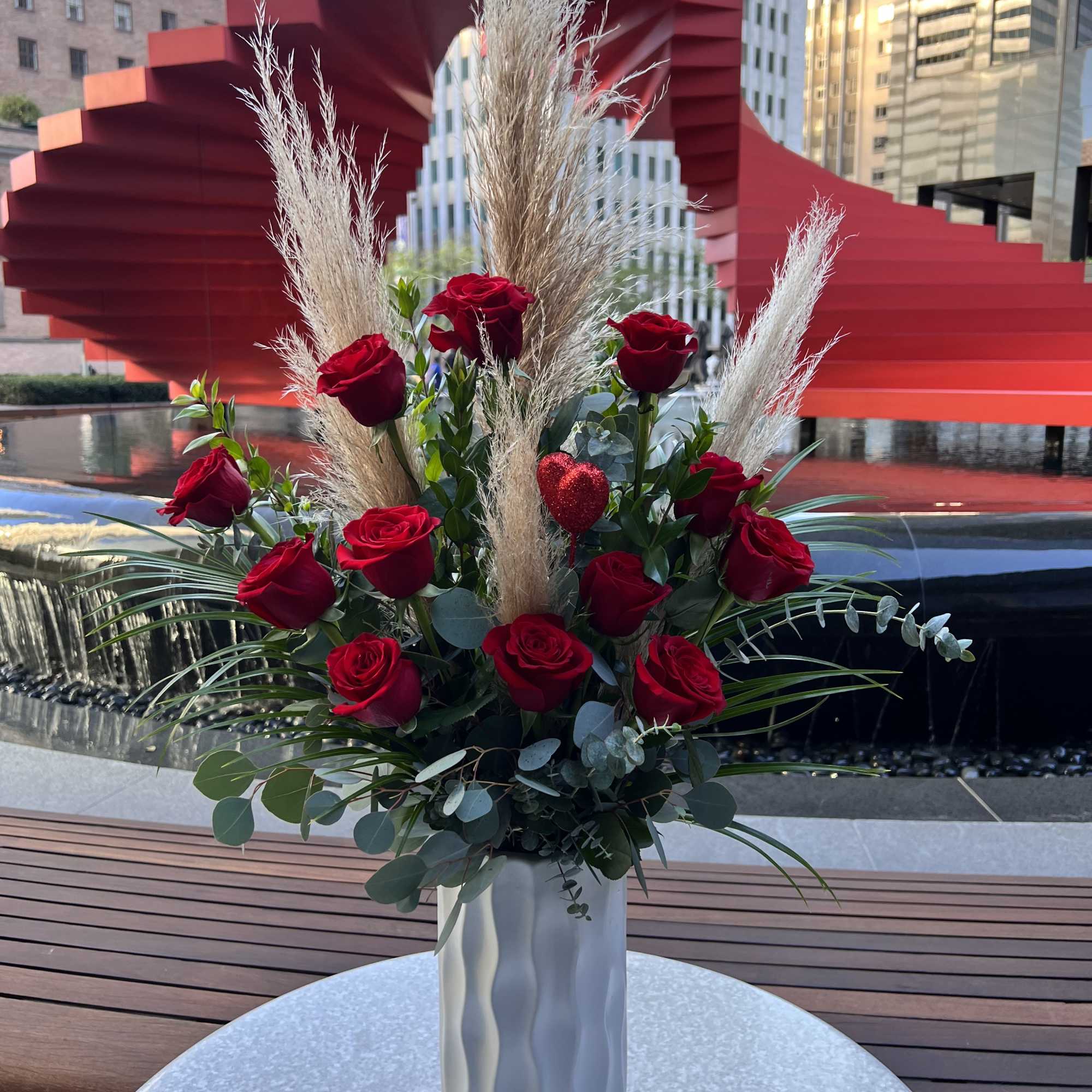 Red roses arranged in a tall white vase with dried grasses.
