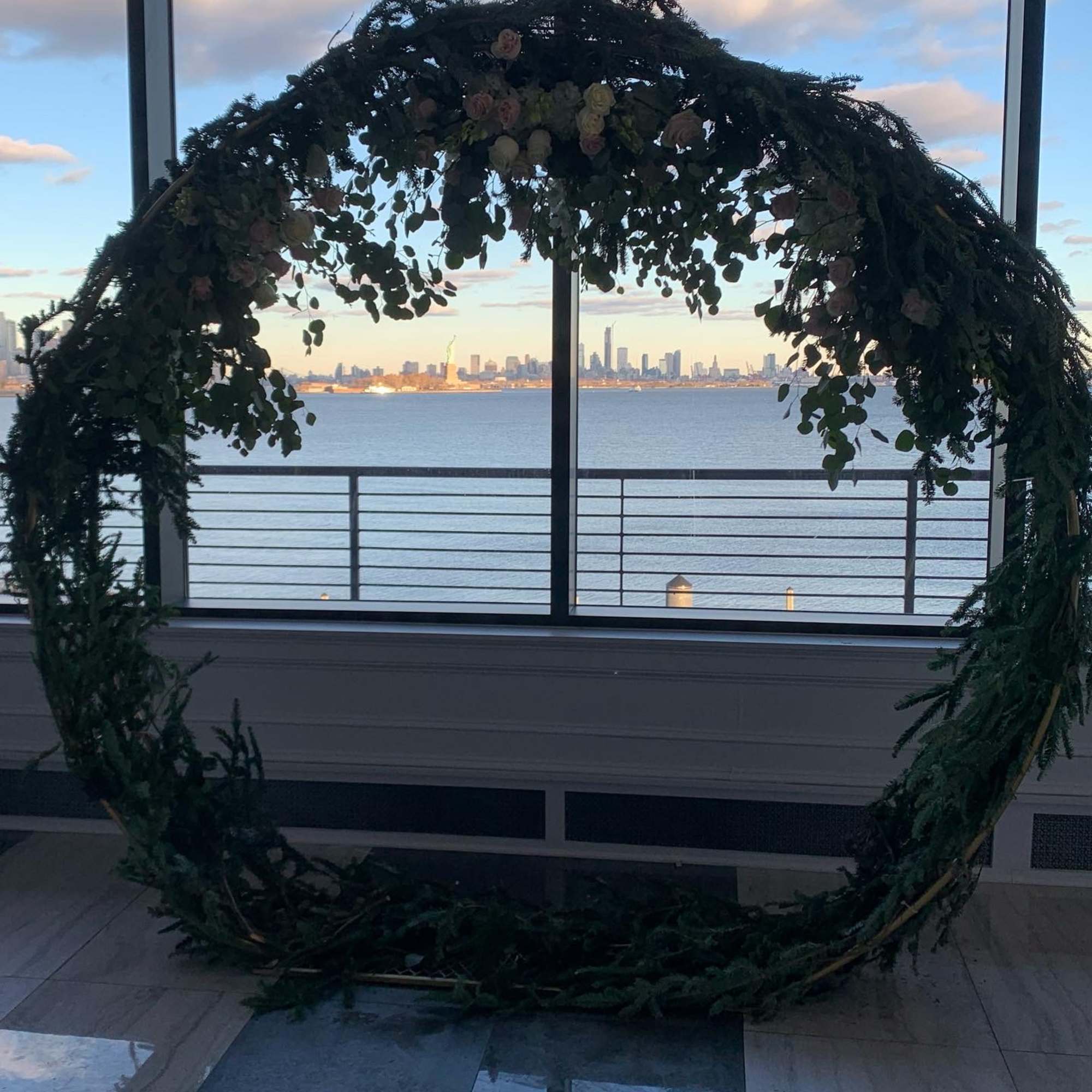 Large circular floral arch with greenery and pale roses by a waterfront window