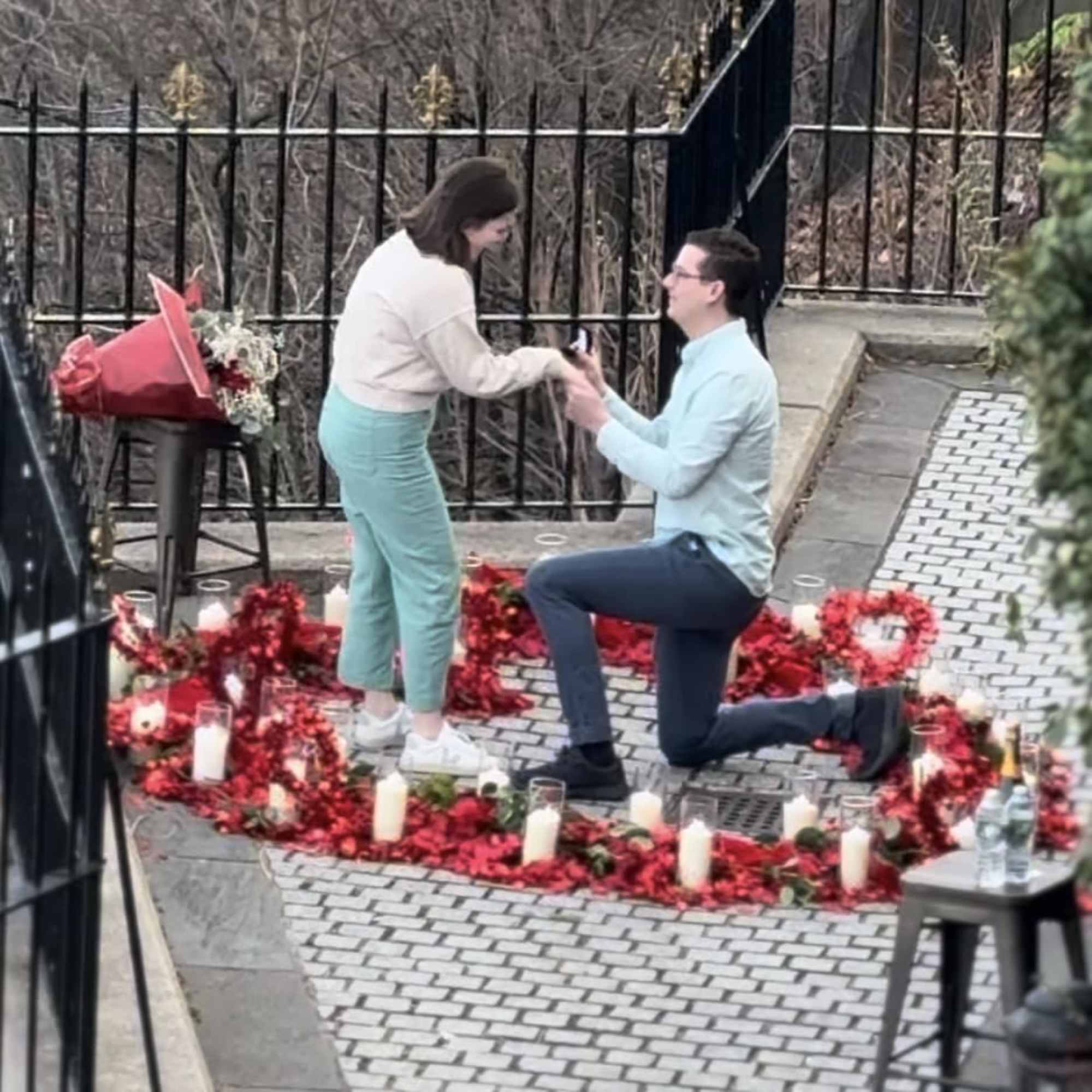 Couple kneeling in a heart-shaped rose petal display with candles and a bouquet nearby
