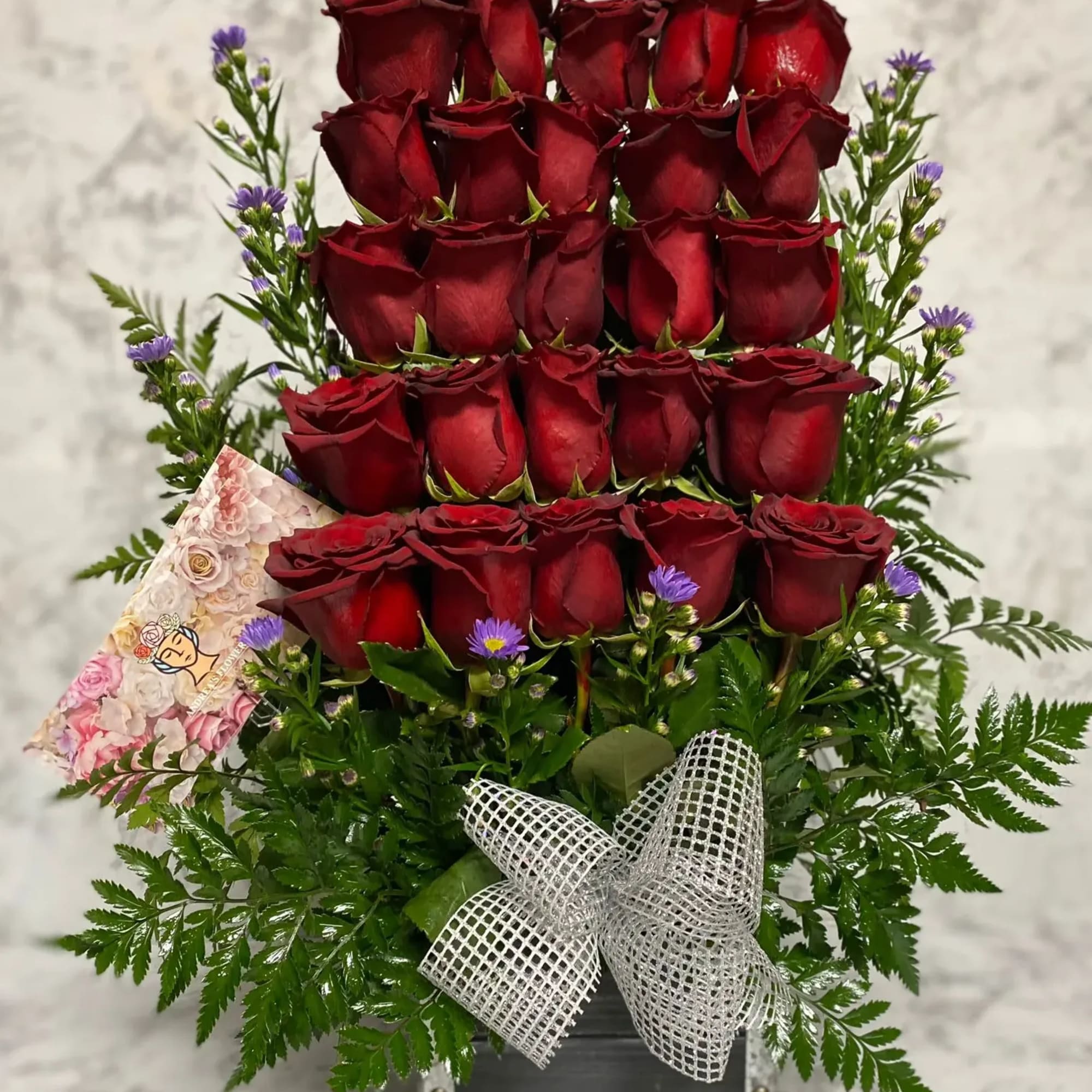 Tall arrangement of tiered red roses with small purple asters in a metal container with a silver bow