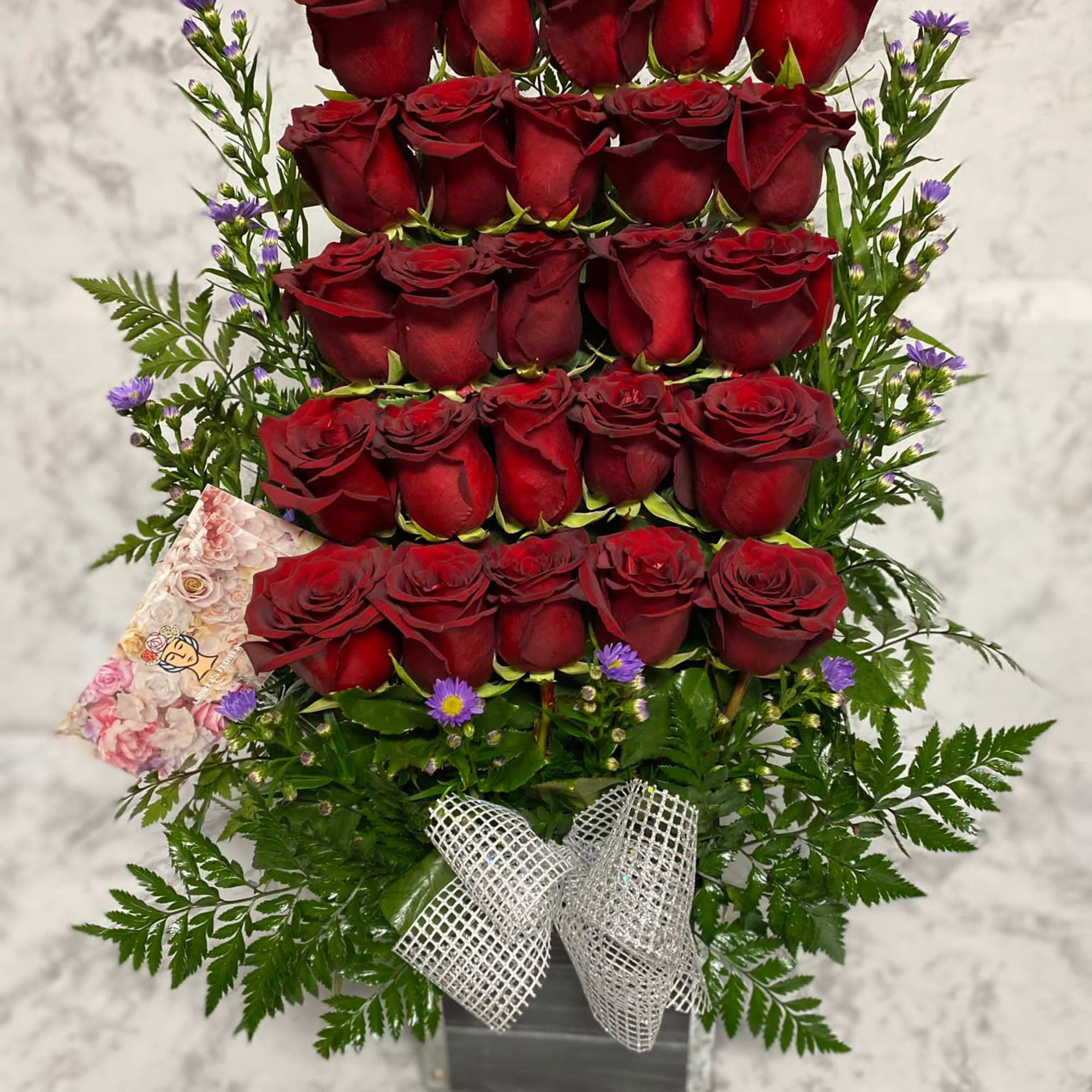 Tall arrangement of stacked red roses with small purple flowers in a black box with a white mesh bow.