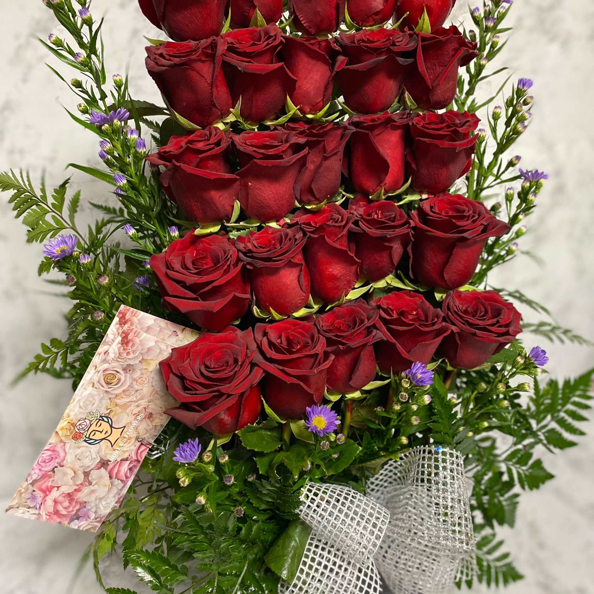Tall tiered arrangement of red roses with small purple blooms in a square container with a silver bow.