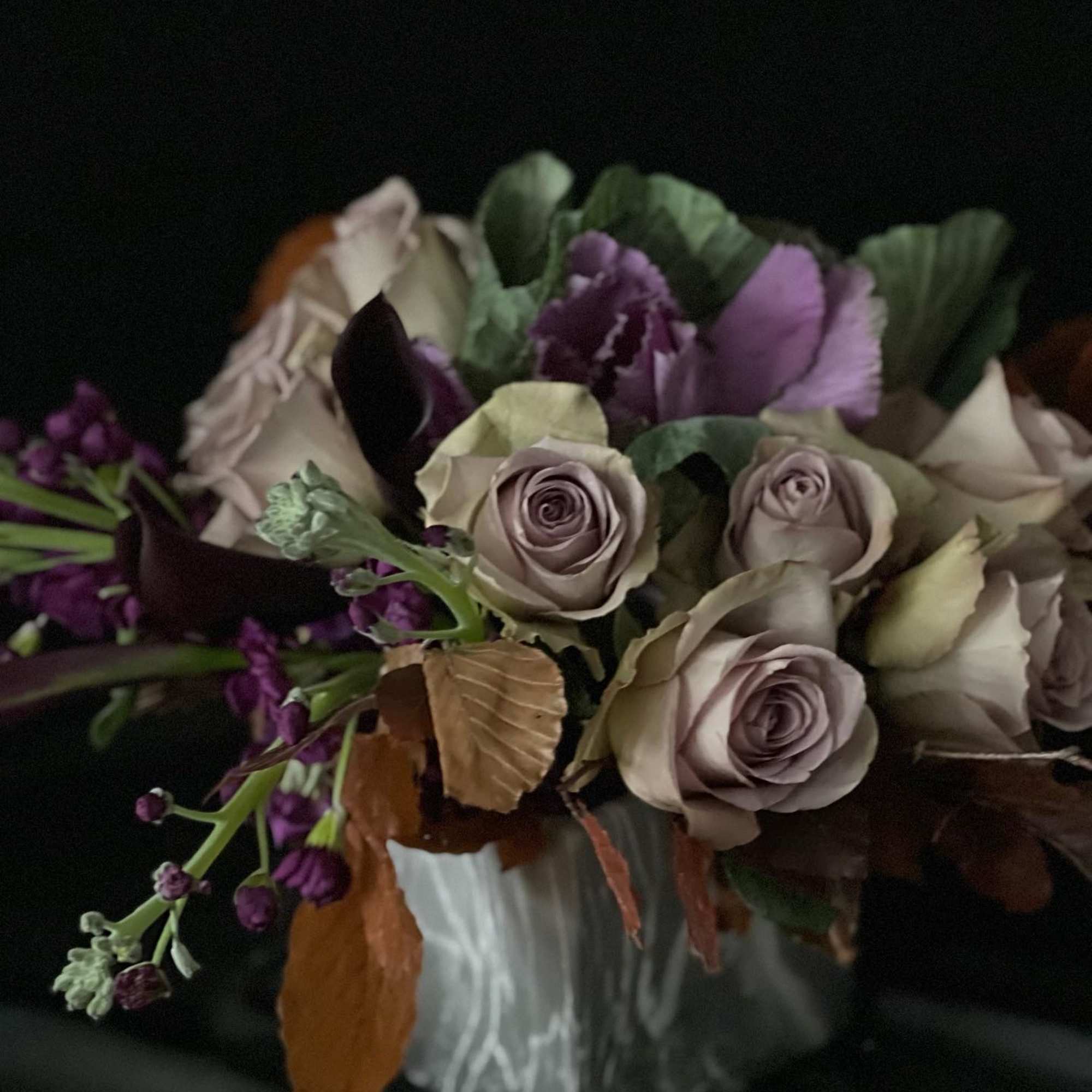 Moody arrangement of lavender roses, dark purple flowers, and kale in a white ceramic vase