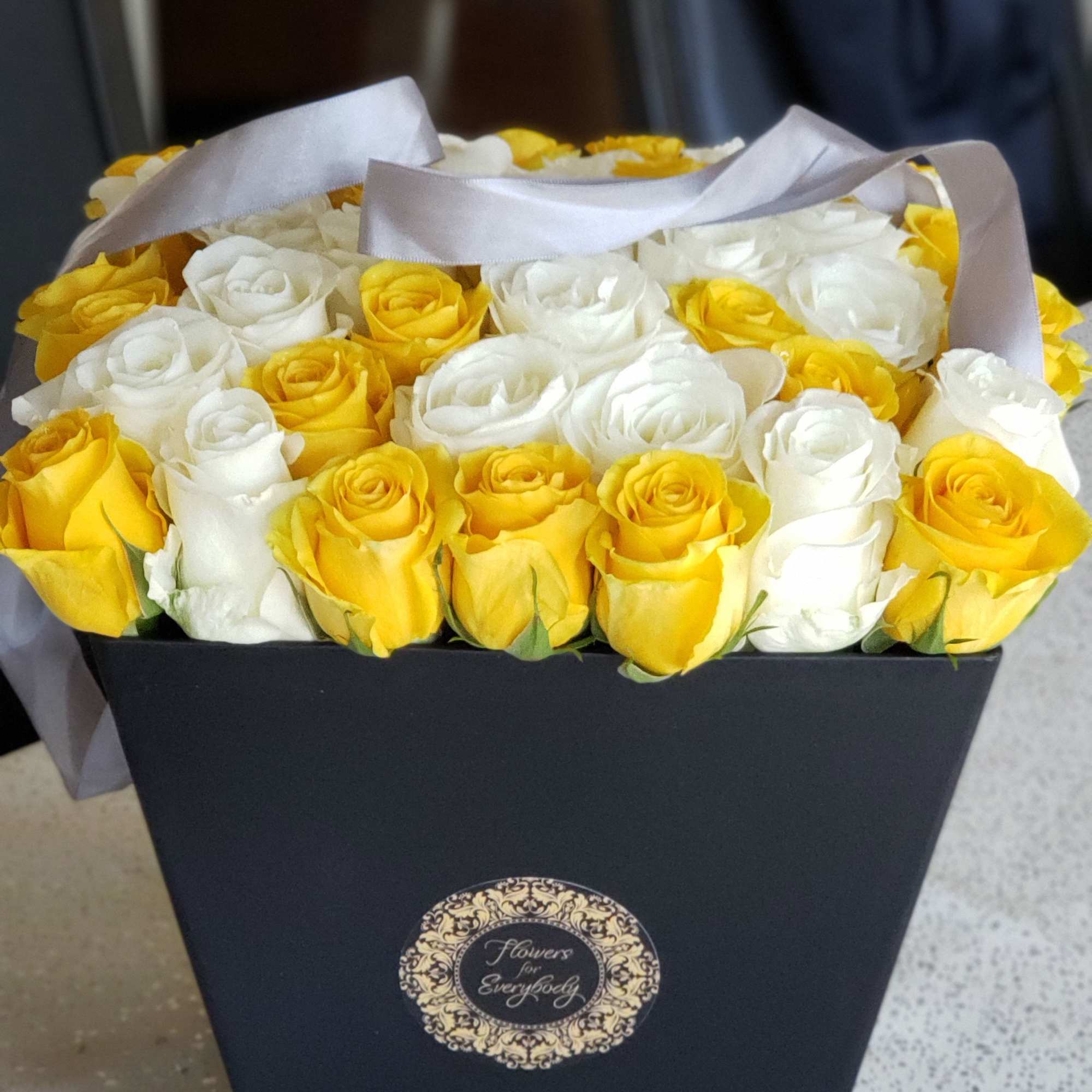 Yellow and white roses arranged in a black box with ribbon handles