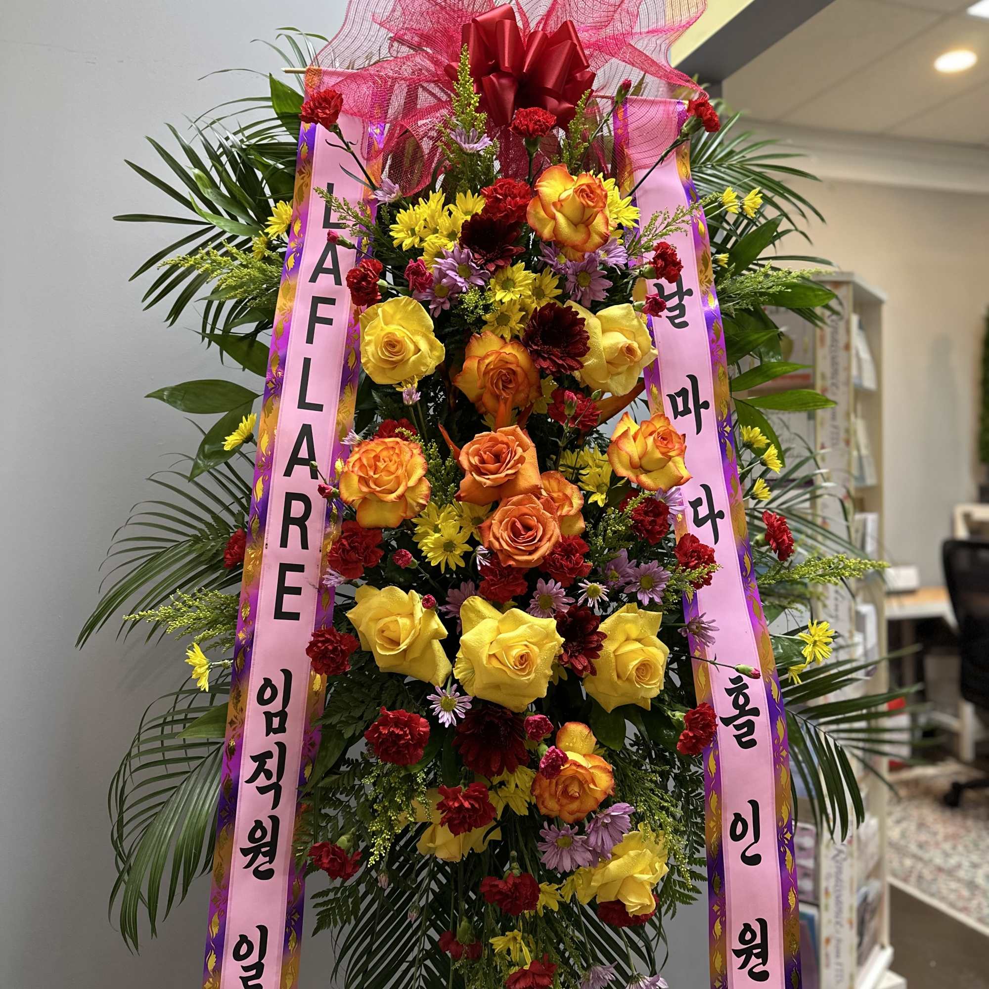 Tall standing floral spray with roses and mixed blooms, topped with a pink ribbon bow