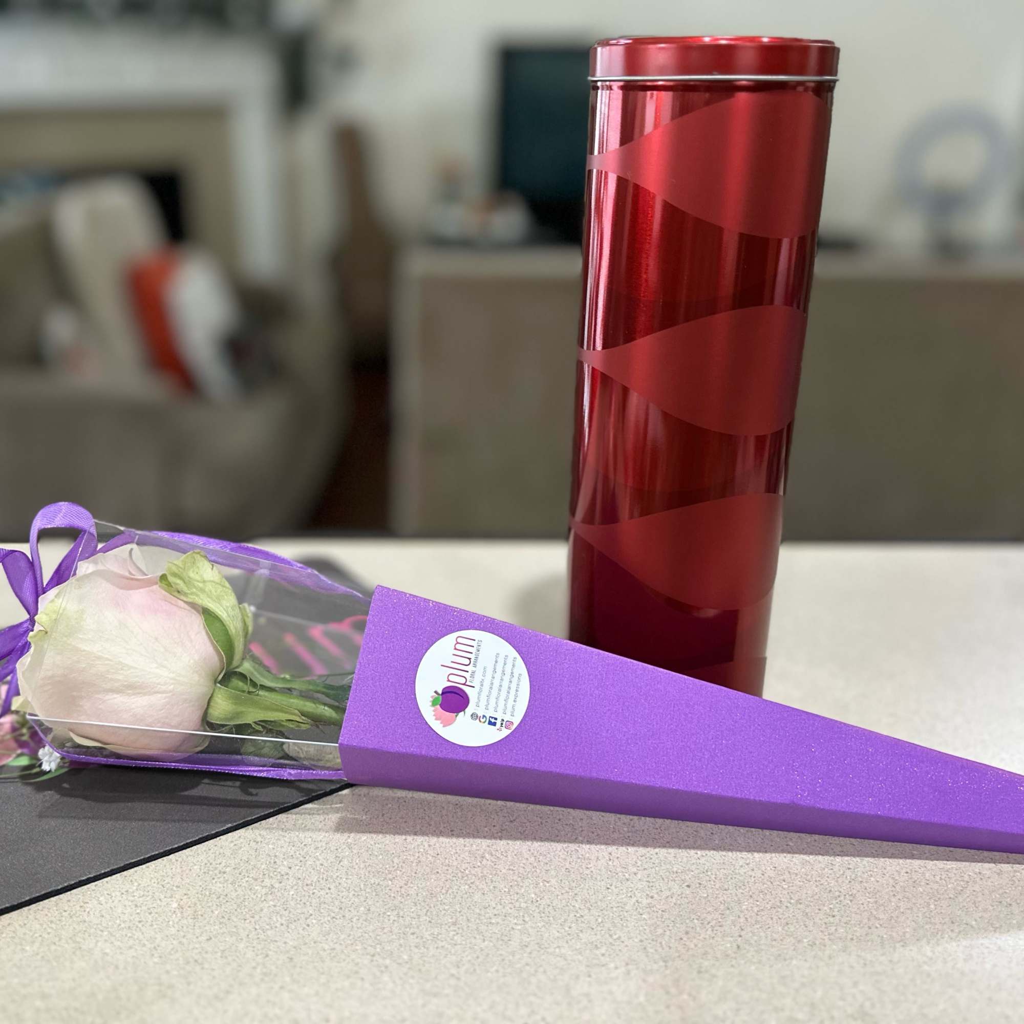 Single pale pink rose in a purple cone wrap beside a red cylindrical gift box
