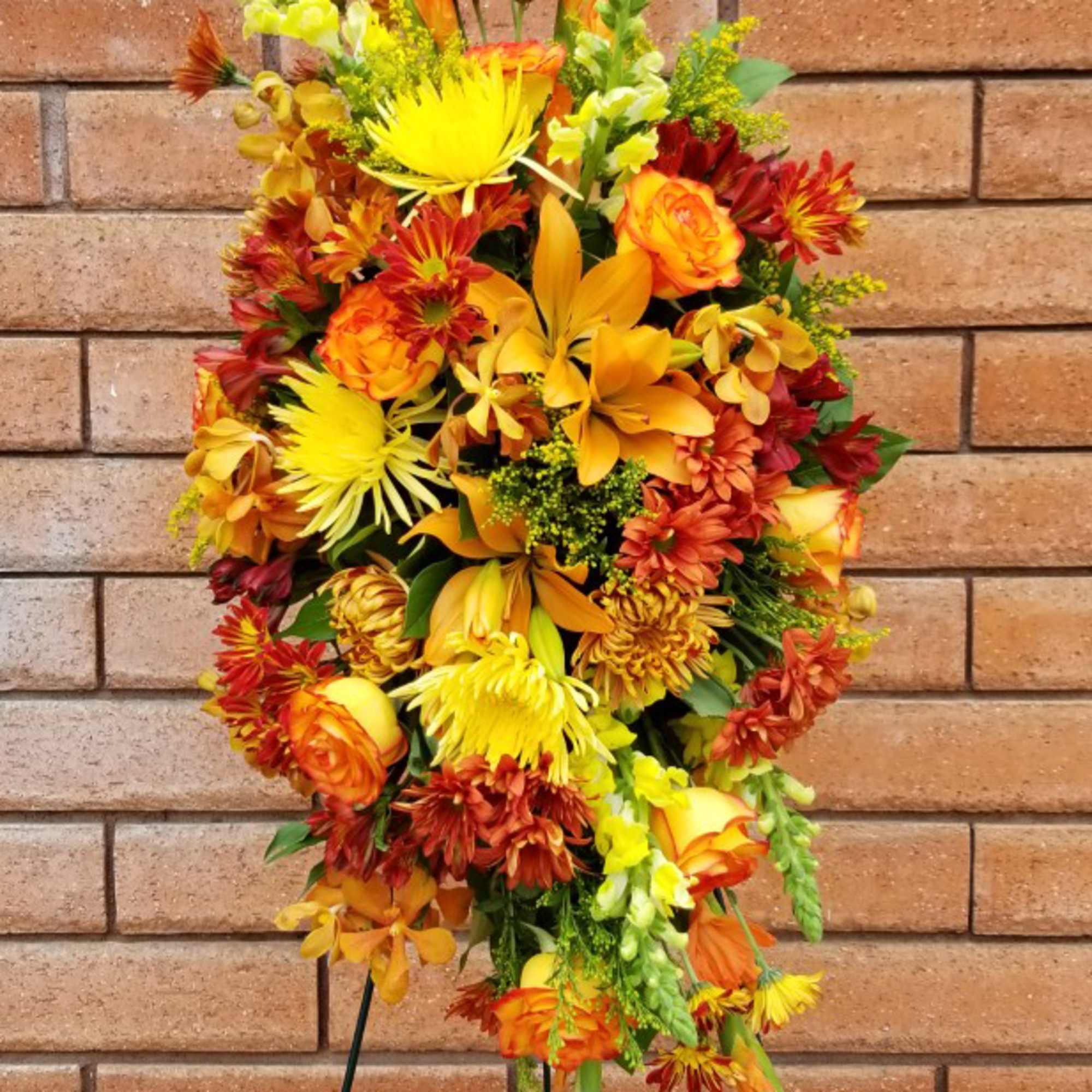 Orange and yellow floral standing spray with mixed blooms against a brick wall