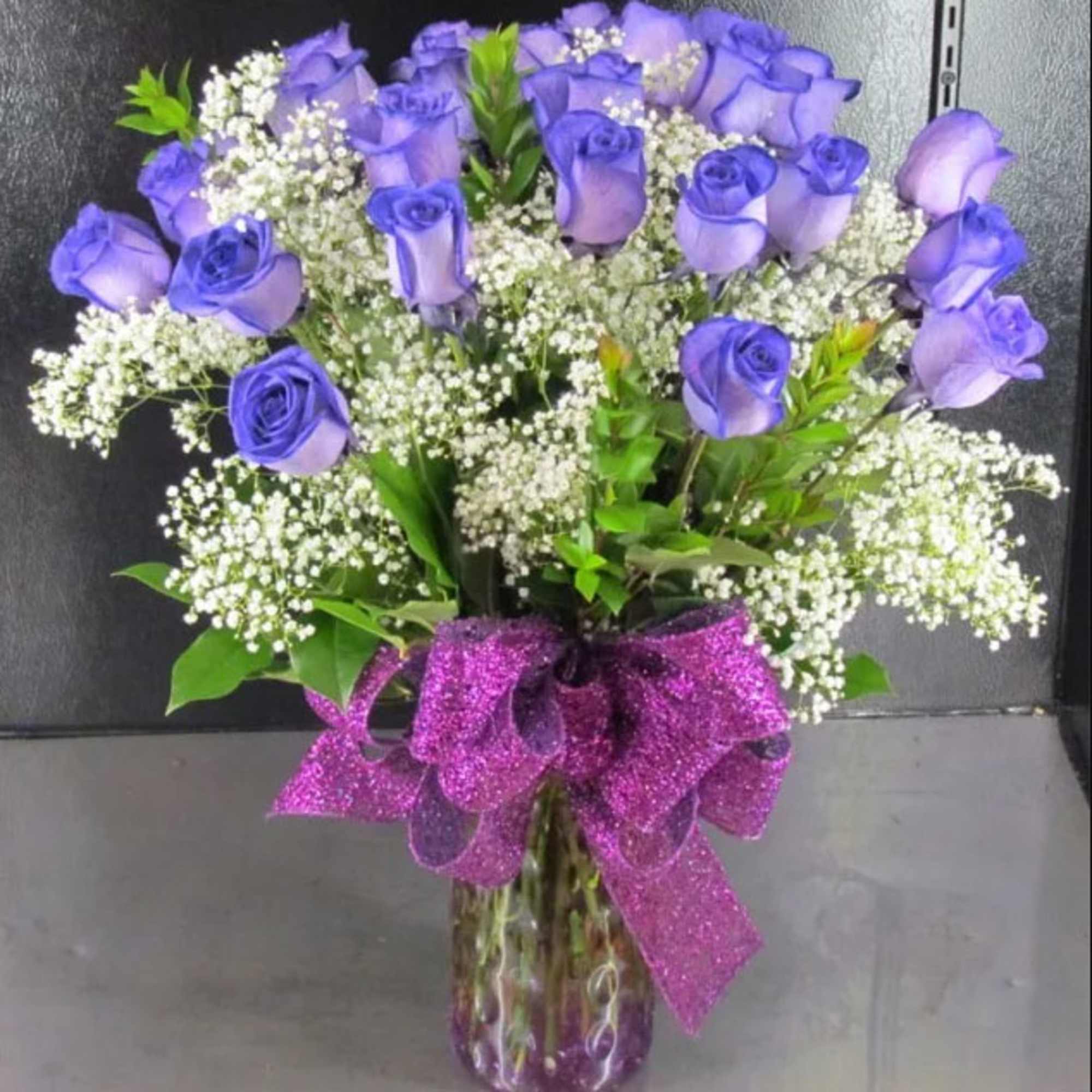 Purple roses in a glass vase with white filler flowers and a glittery bow
