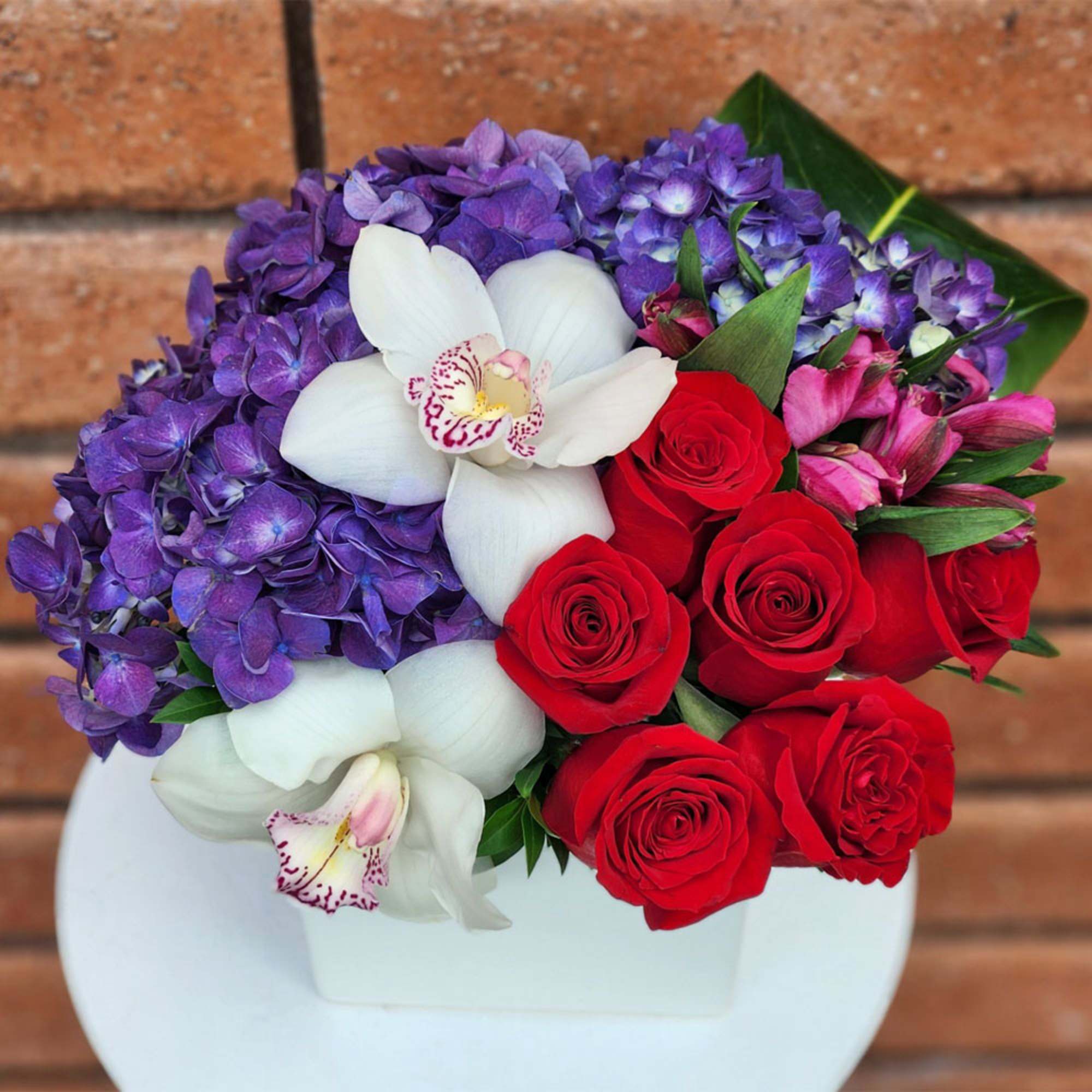 Bouquet of red roses, purple hydrangeas, and white orchids in a white container