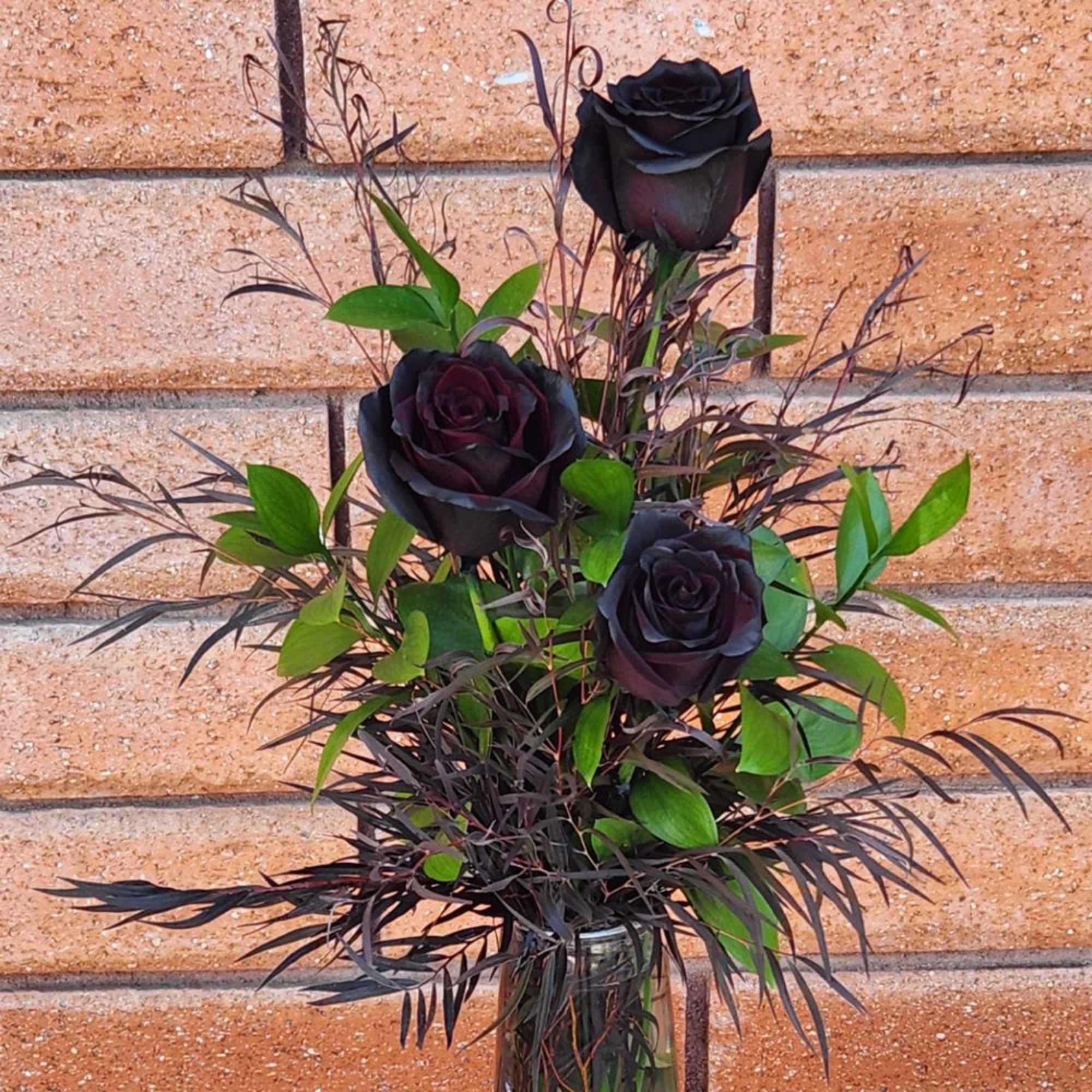 Arrangement of three black roses with greenery in a clear glass vase