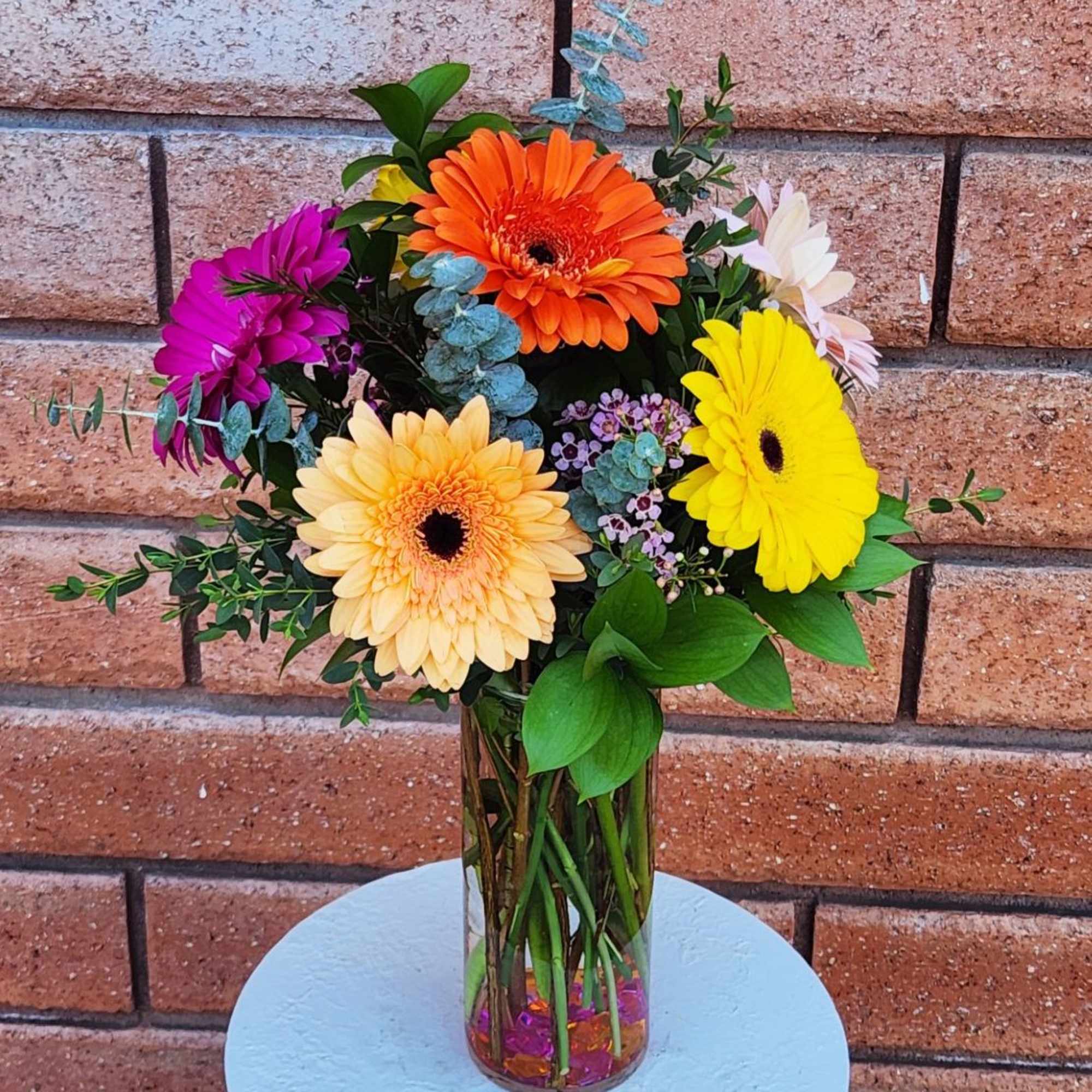 Tall glass vase with bright gerbera daisies in orange, yellow, peach, and magenta with filler blooms.