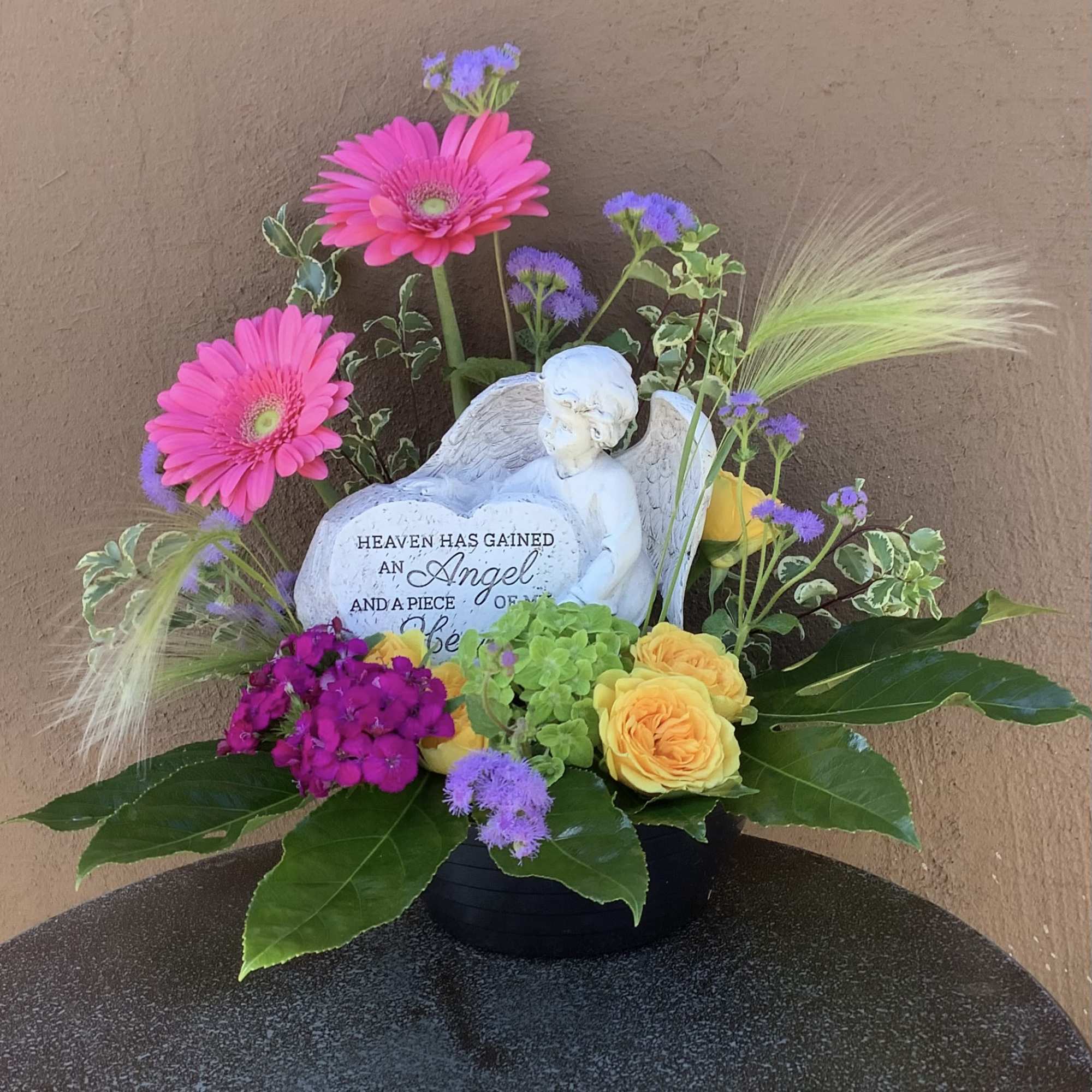 Floral sympathy arrangement with pink gerbera daisies and yellow roses around an angel figurine