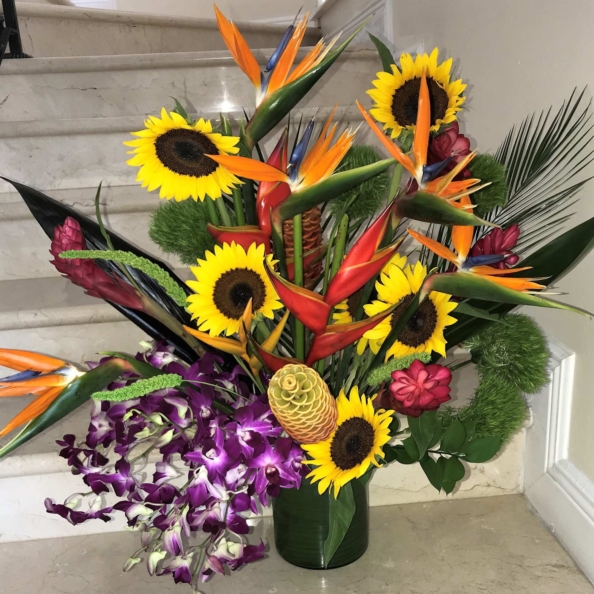 Tall tropical arrangement with sunflowers, birds of paradise, heliconia, and purple orchids in a glass vase