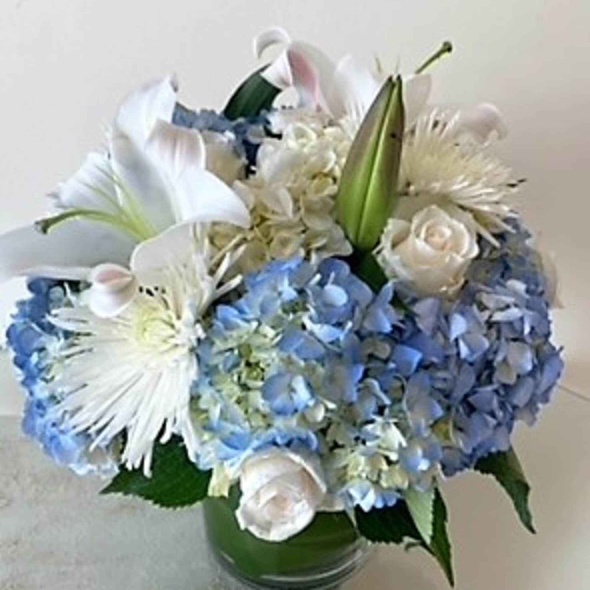 Round arrangement of blue hydrangeas, white lilies, roses, and mums in a clear glass vase