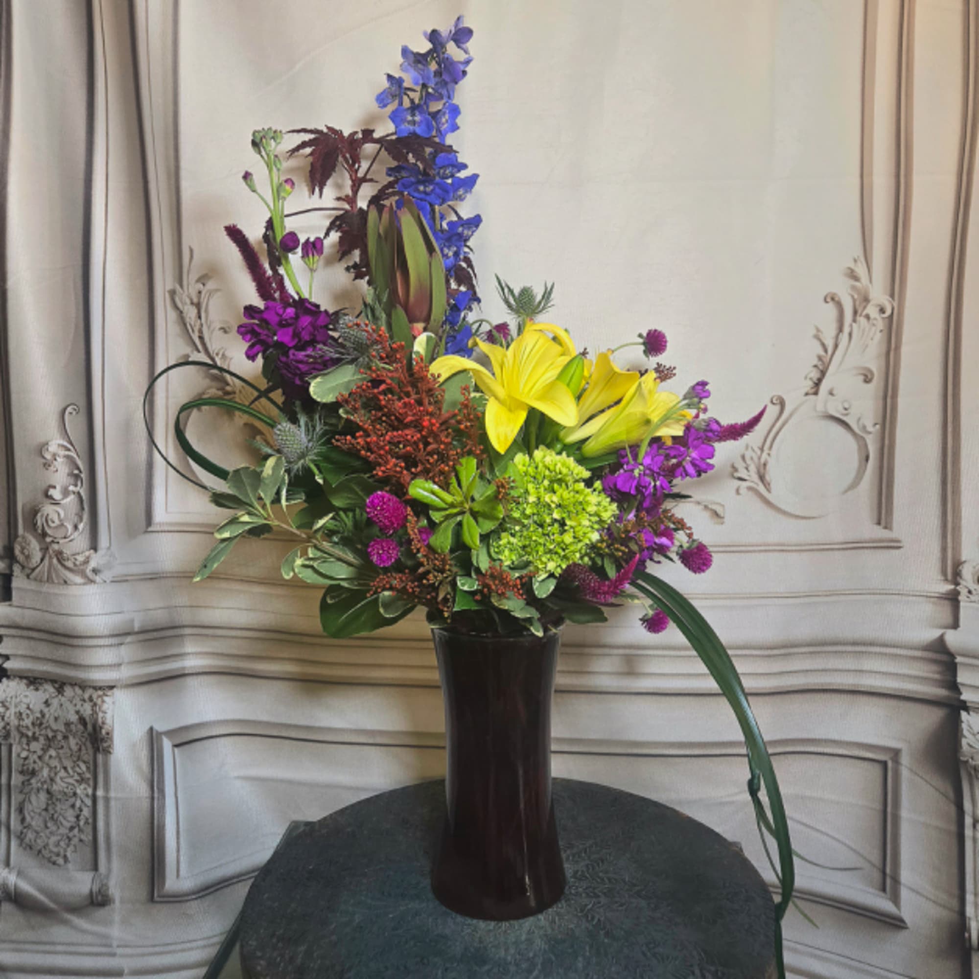Tall mixed flower arrangement in a dark vase with yellow lilies and purple blooms
