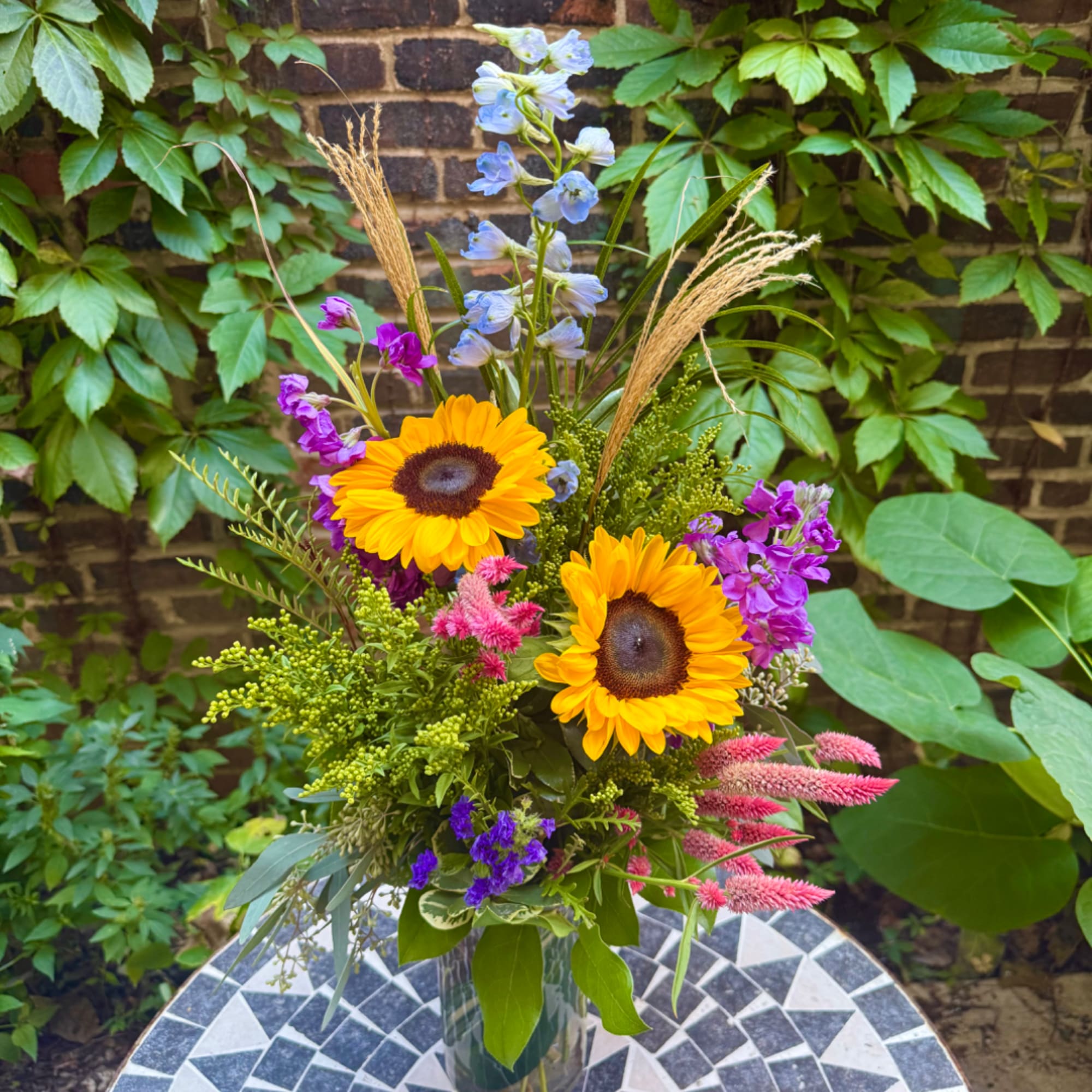 Mixed bouquet with sunflowers, blue delphinium, and pink accents in a glass vase